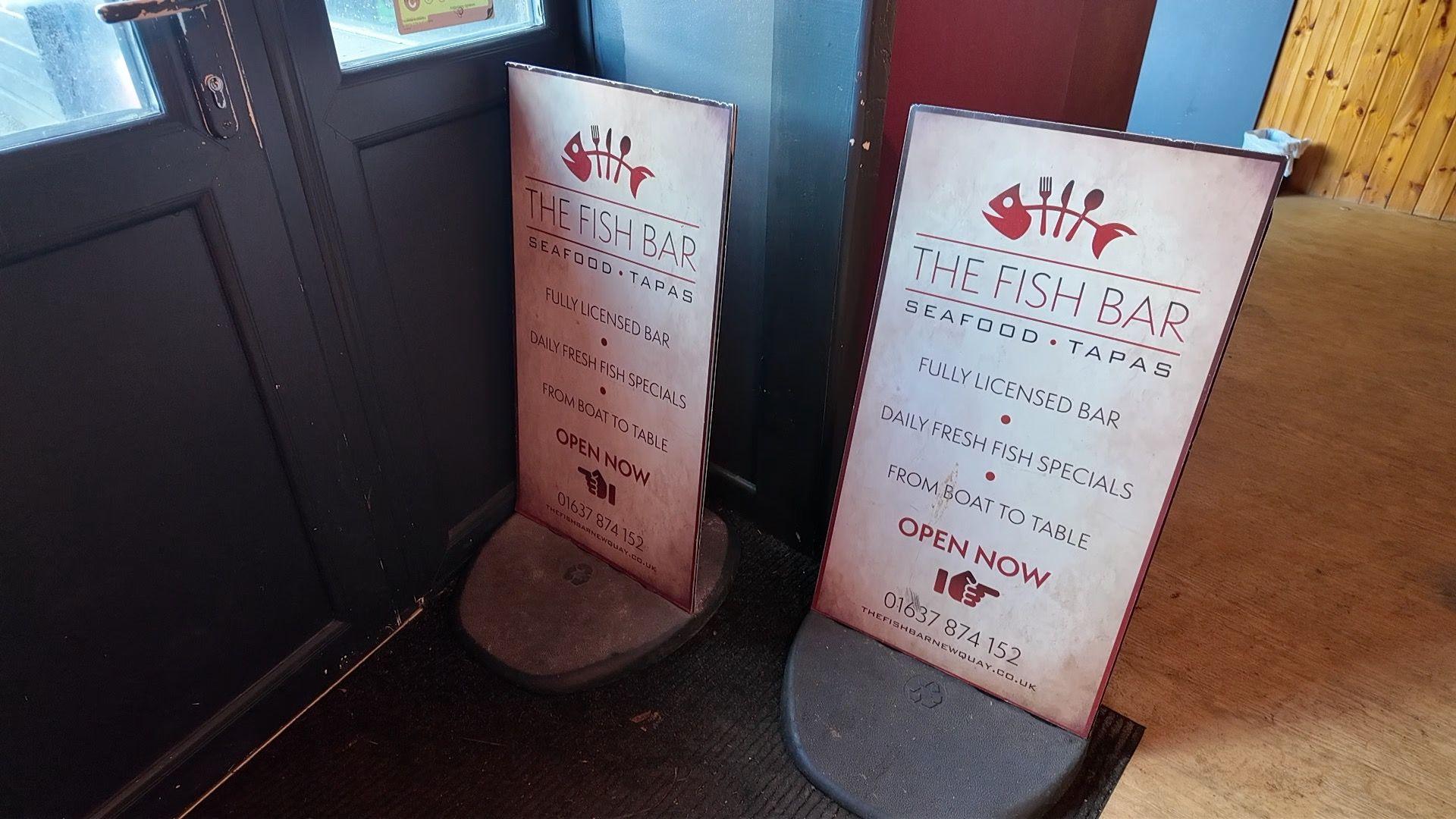 A close-up image of two, street-side restaurant signs that are inside the front door of a restaurant. They say The Fish Bat, Seafood, Tapas.