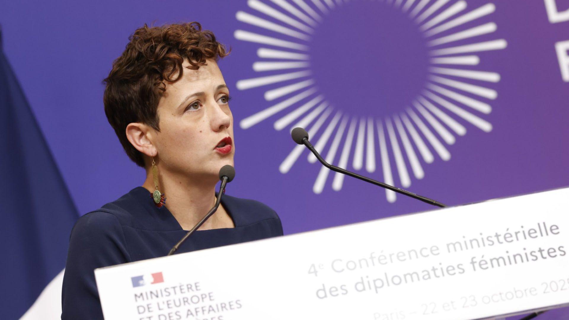 A woman with short, dark, curly hair speaks at a conference. She is wearing a navy blue top with dangly earrings. A podium with two microphones is in front of her and a purple screen is behind her.