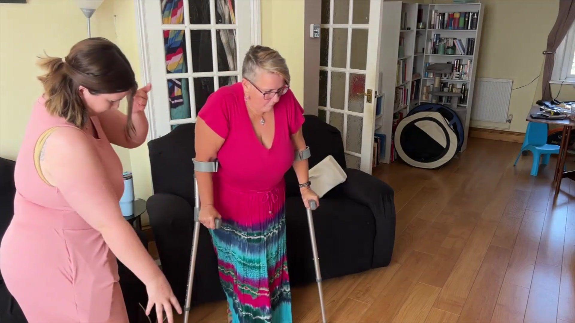 Jenefer Sully, wearing a pink shirt and tie-dye skirt is using crutches to get up from a sofa, whilst a family member - wearing a pink dress - helps her up