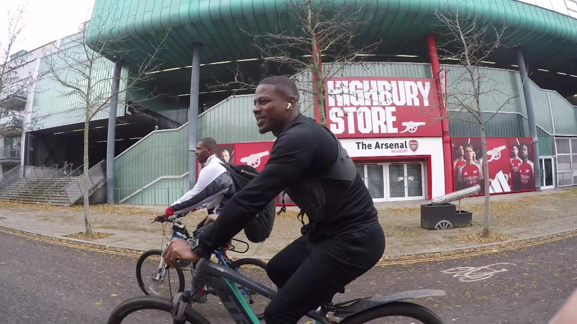 Two men on bikes cycling past the Arsenel stadium. In the background there is the Arsenal shop with red and white shop sign saying Highbury Store and The Arsenal underneath.