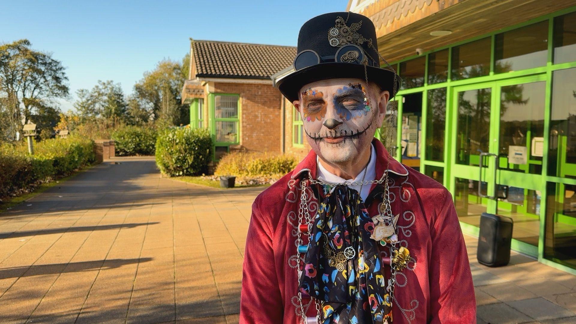 Principal Seán Dillon stands outside the school, he is wearing a black top hat with steampunk style flying goggles on it, white make up with black detailing round the mouth, a velvet red coat and a multicoloured silk cravat/scarf. 