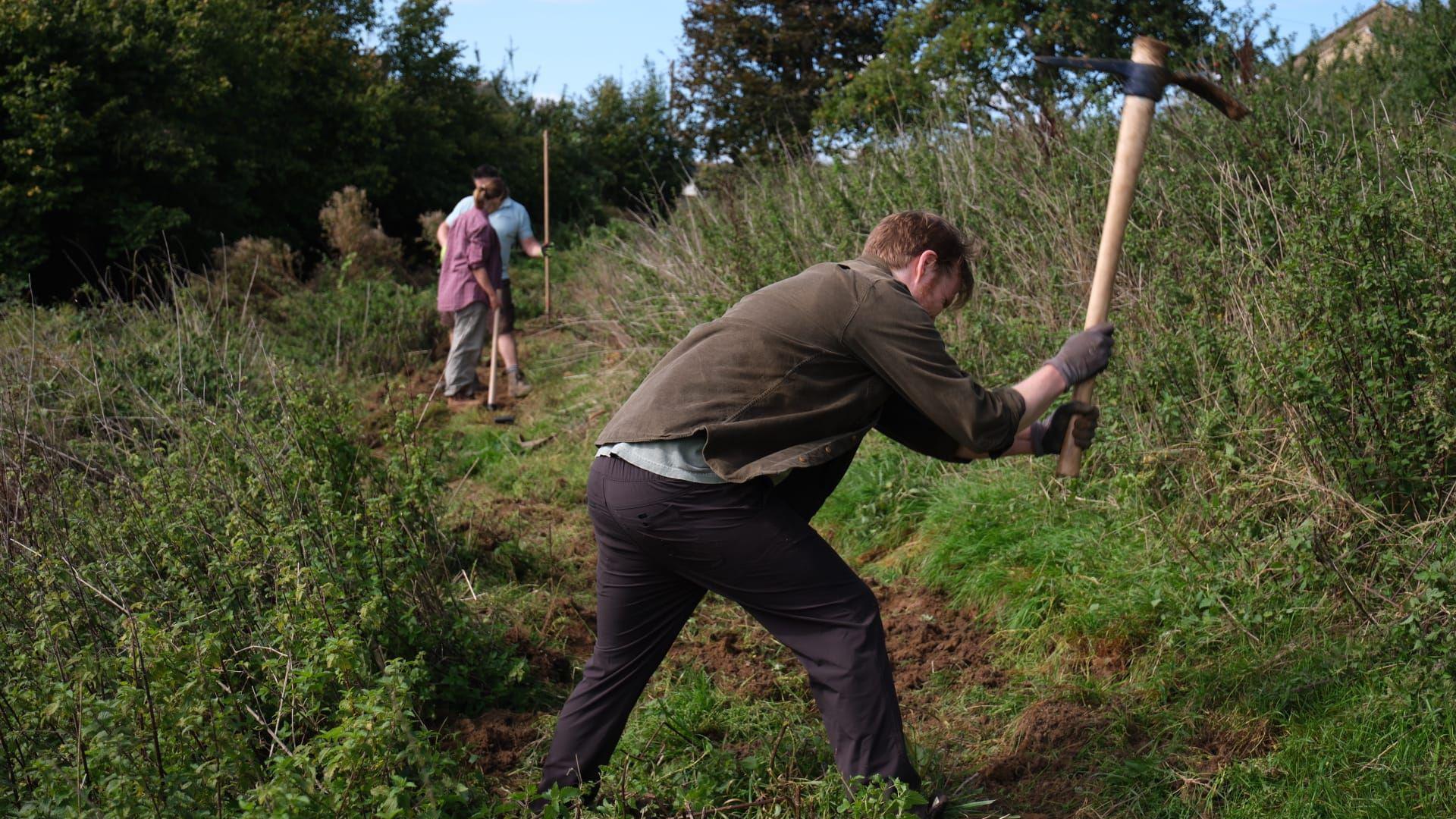 A man with light hair takes a pick to the soil on a slope at Heavens Valley near Stroud, surrounded by nettles. Two other people - a man and a woman - also with tools can be seen standing a few metres away from him.
