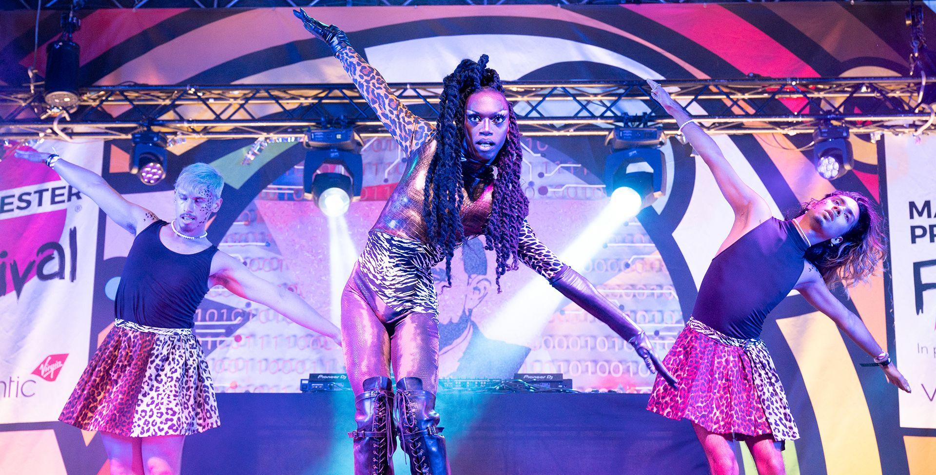 Artist Black Peppa performs during Manchester Pride 2022 - they're pictured mid-dance with their arms outstretched while wearing leopard and zebra print spandex alongside two performers wearing leotards.