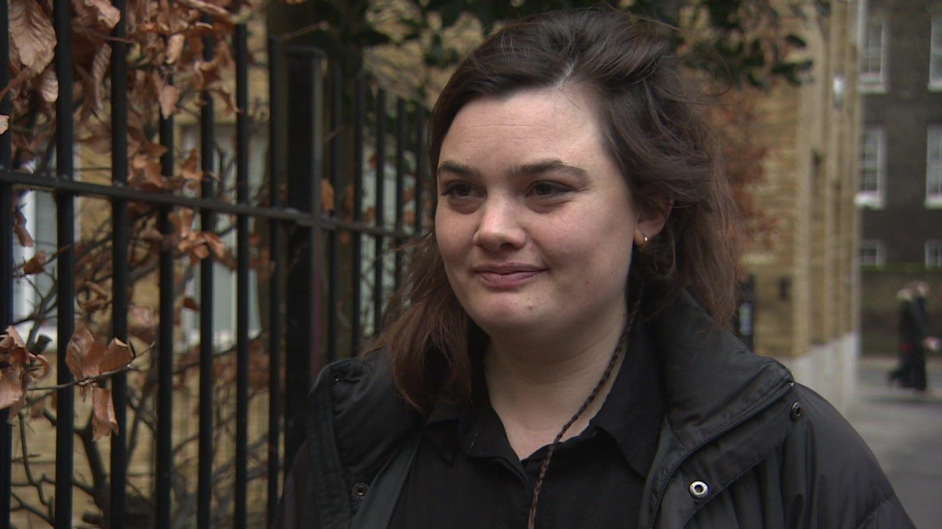 Bekah Sparrow, a young woman with long dark hair wearing a black shirt and black coat, looking at the reporter to the side of the camera. Black railings with branches and brown leaves can be seen in the background