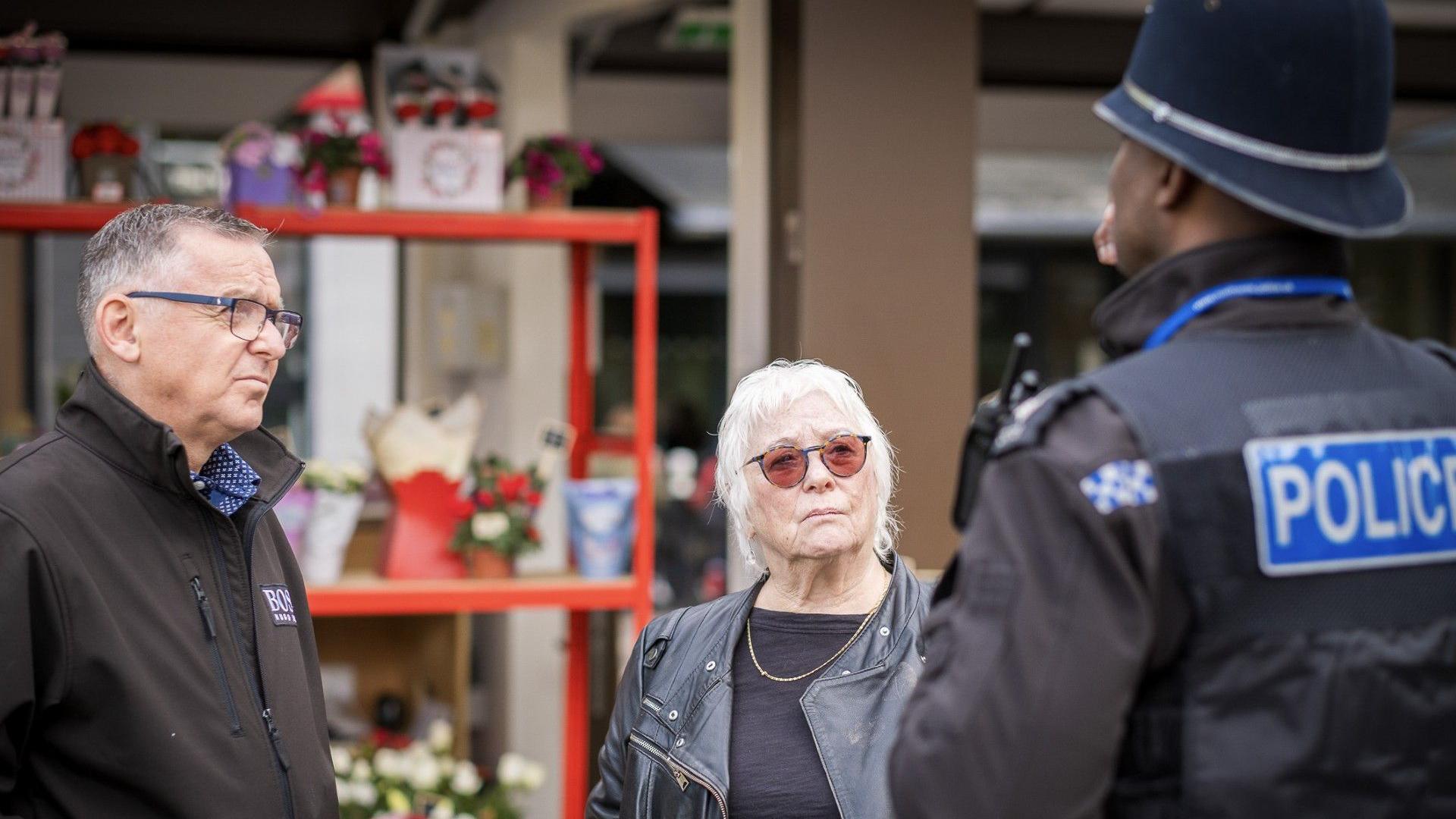 Danielle Stone (middle) standing by a man, wearing a black jacket and a police officer to the right. She has on glasses, a black leather jacket and a black top. She is looking at the police officer.