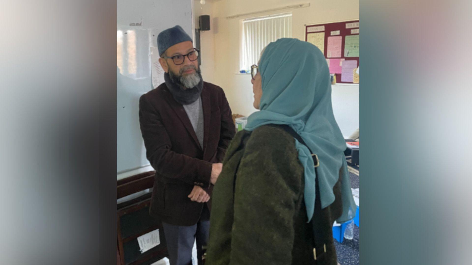 A man wearing a blue skull cap, with glasses and a grey beard, is shaking hands with a woman wearing a blue scarf over her head and a dark green coat. They are in a training room with white walls and a noticeboard.