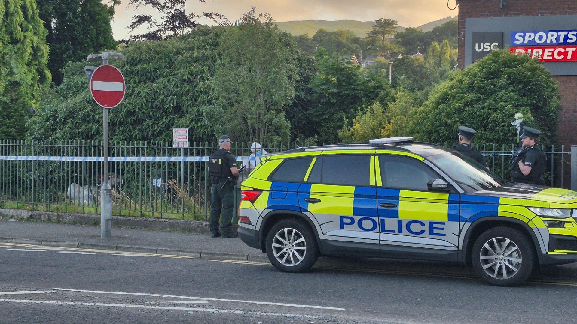 A yellow and blue police car sits half on the road, half on the footpath. There are three police officers behind the car dressed in green trousers and t-shirts with black body armour vest over them. A blue and white cordon is in place behind the men. A man in all white overalls and blue mask stands behind the cordon in a green bushy area.
