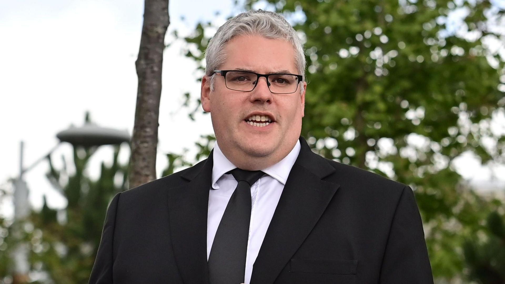 Gavin Robinson has short grey hair and black framed glasses. He is wearing a black suit, dark plain tie and white shirt. Trees are in the background.