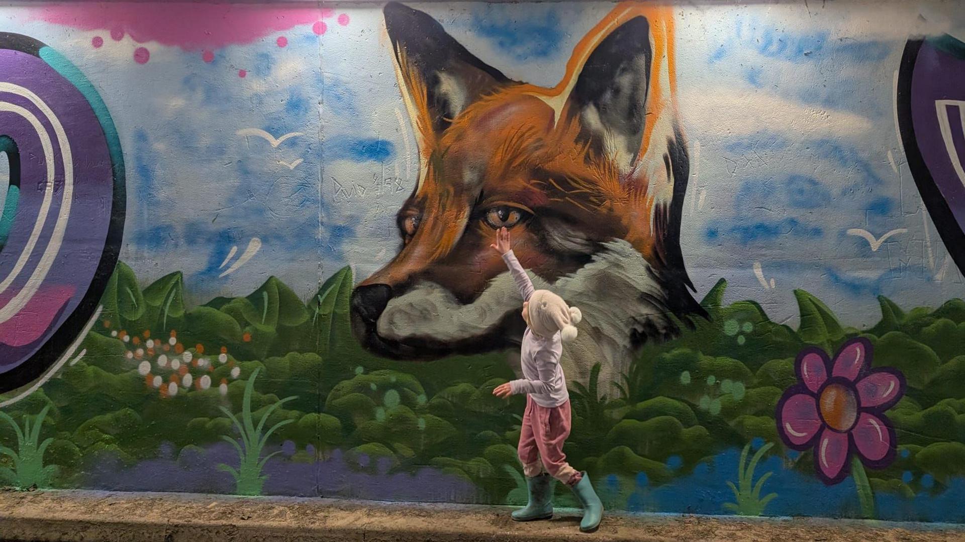 A young girl reaches to touch a painted fox on the subway wall