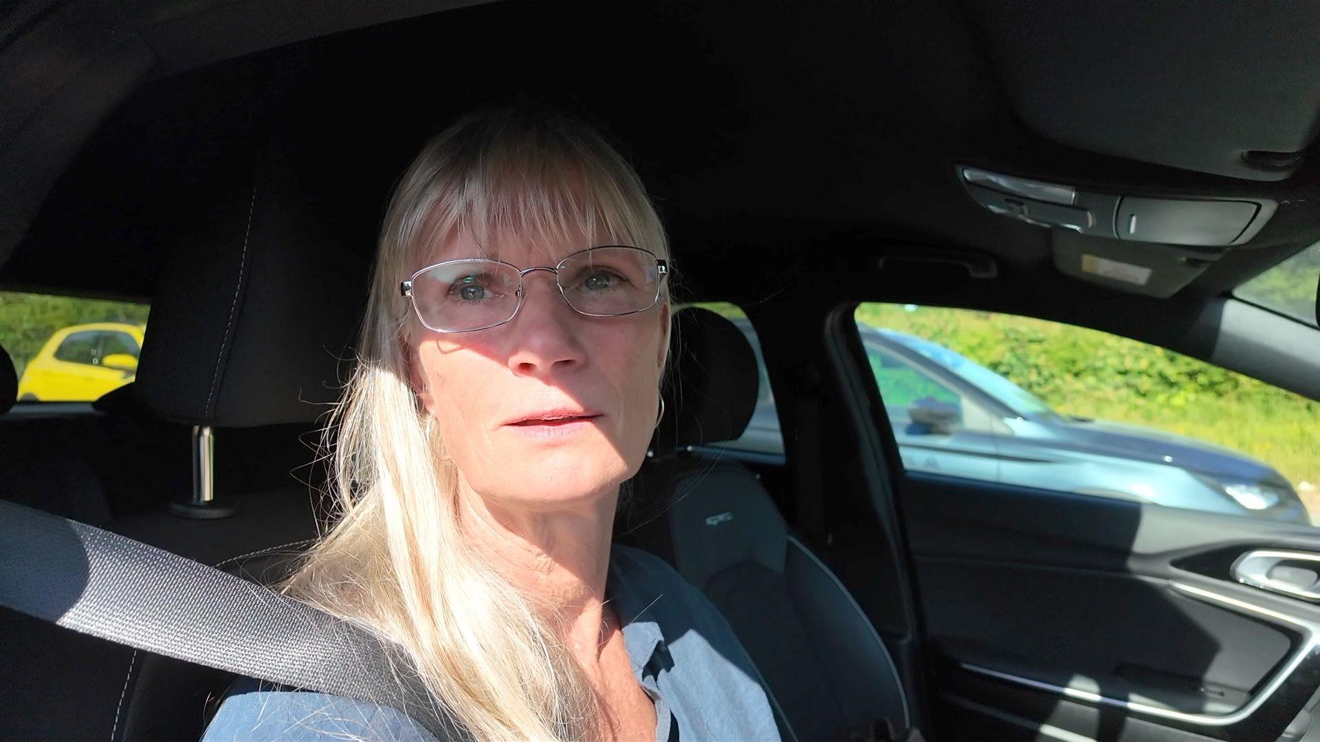 A blonde woman wearing glasses sits in a car frustrated by the delays caused by the rescheduled roadworks.