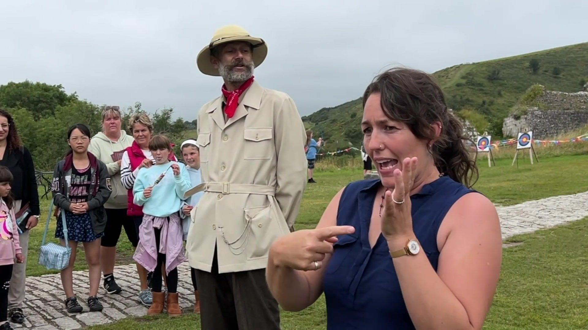 The BSL interpreter translating next to the castle guide and keeper. There are a few people next to them. Green hills can be seen behind them.