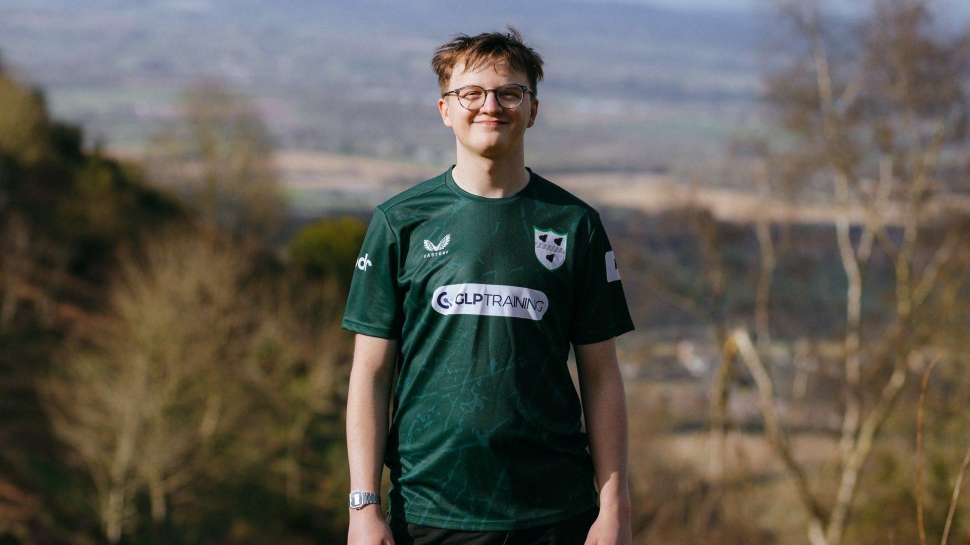 Paddy is outdoors with trees behind him, smiling at the camera. He is wearing glasses and a green football top.