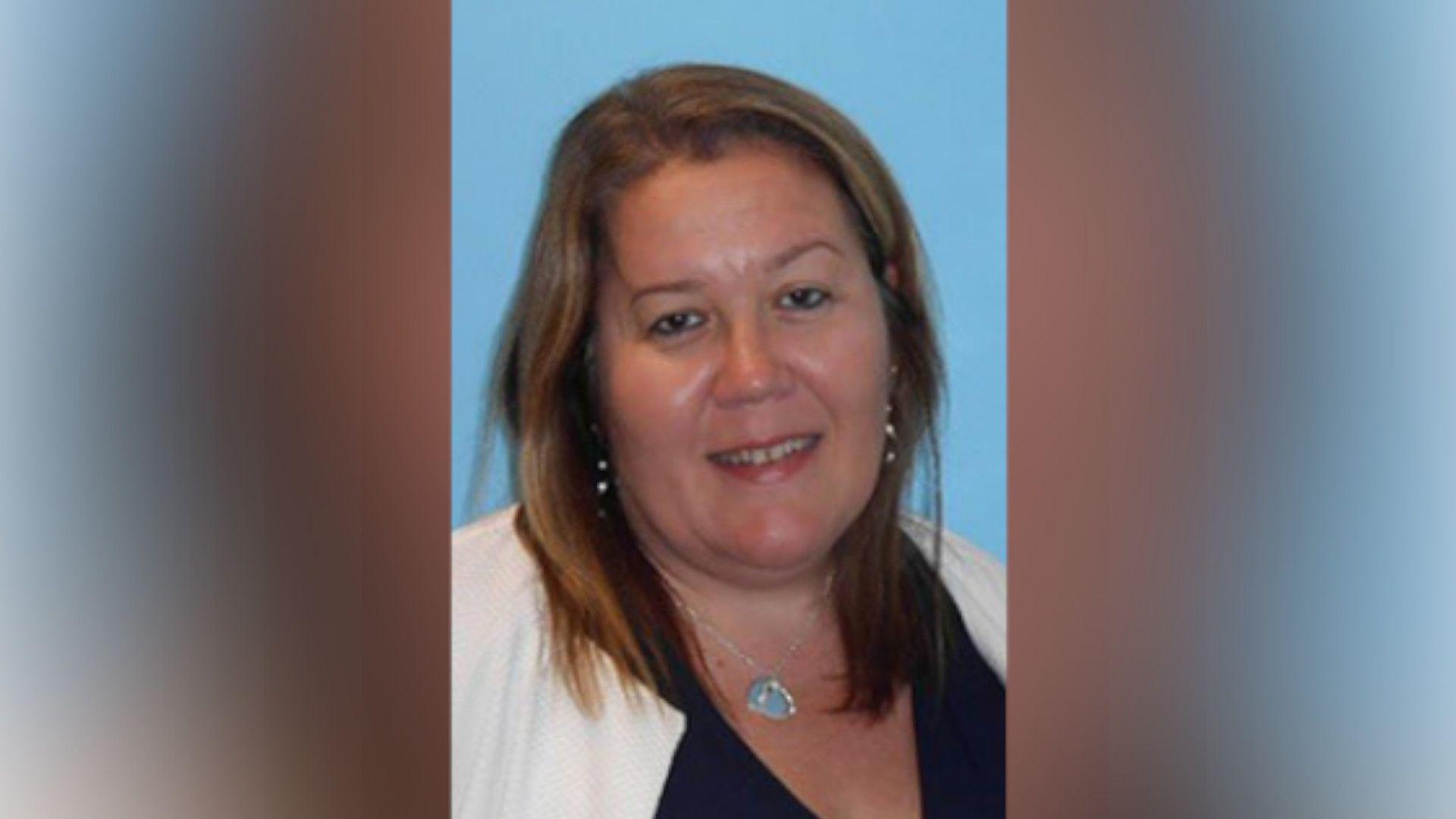 Sarah Gallagher with long brown hair wearing a white jacket and black top. She has a necklace with a small blue triangular charm