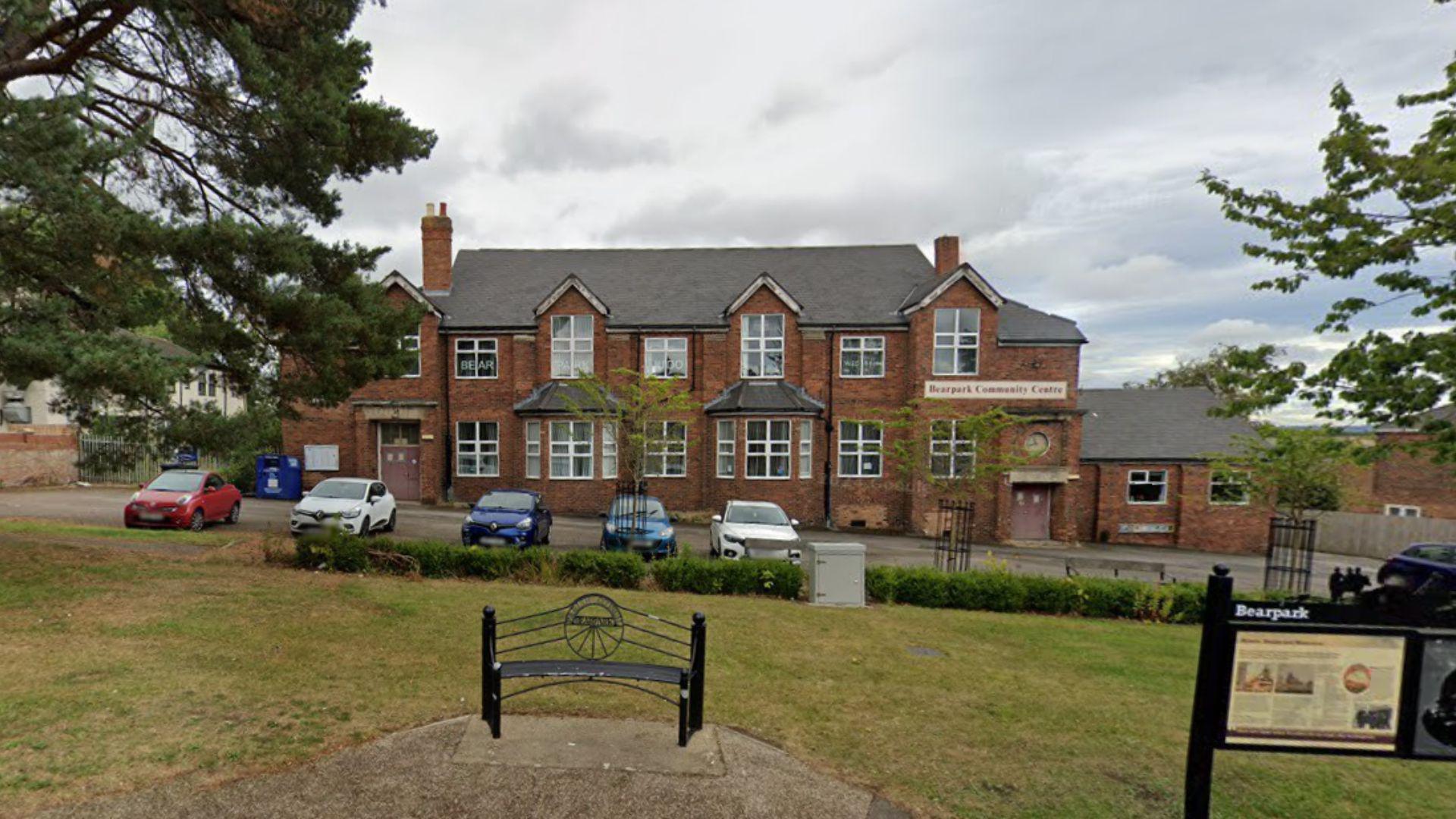 Bearpark Community Centre. It is a three-storey red brick building with a number of windows including two bay windows. Cars are parked outside. It overlooks a park, which includes a park bench and a sign explaining the history of Bearpark.