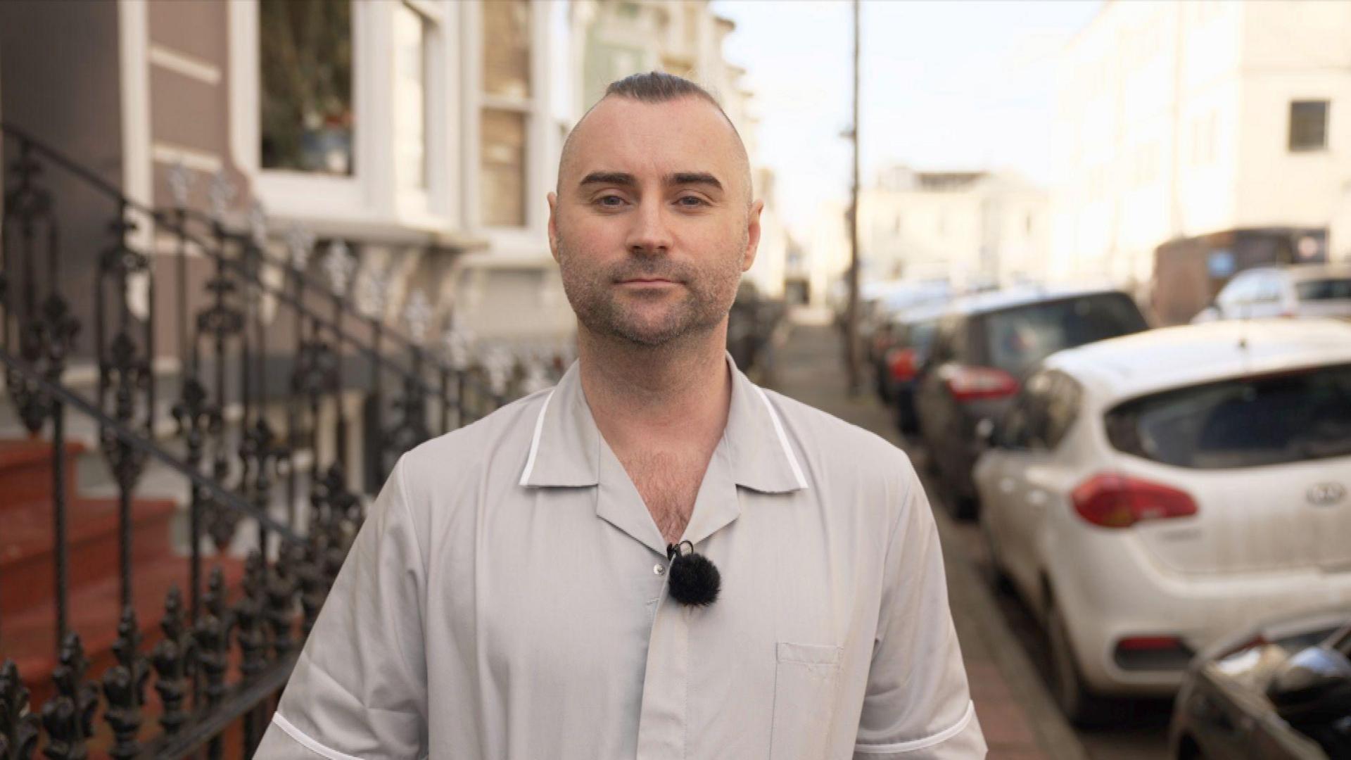 Vaughan Riley, wearing a set of grey healthcare workers' scrubs, stands in a Brighton street, with a row of terraced houses and a parked cars behind him.