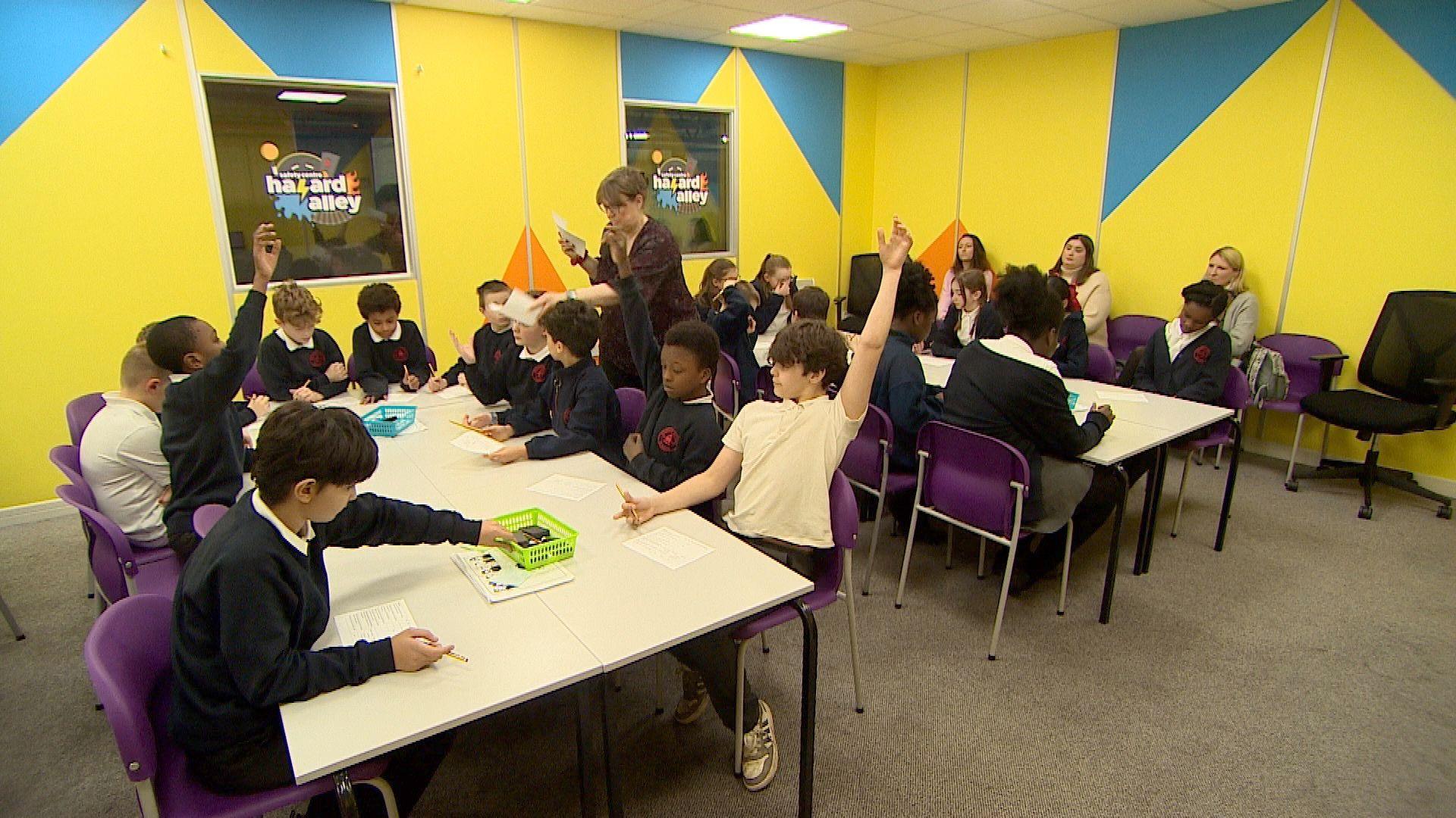 A group of 20 schoolchildren, all aged between 10 and 11, are sat on purple chairs at white desks. Some of them have their hands in the air as they look to get the attention of the course leader who is handing out bits of paper. Watching on is a teacher and two teaching assistants. They're all sat in a bright yellow room with grey carpet.