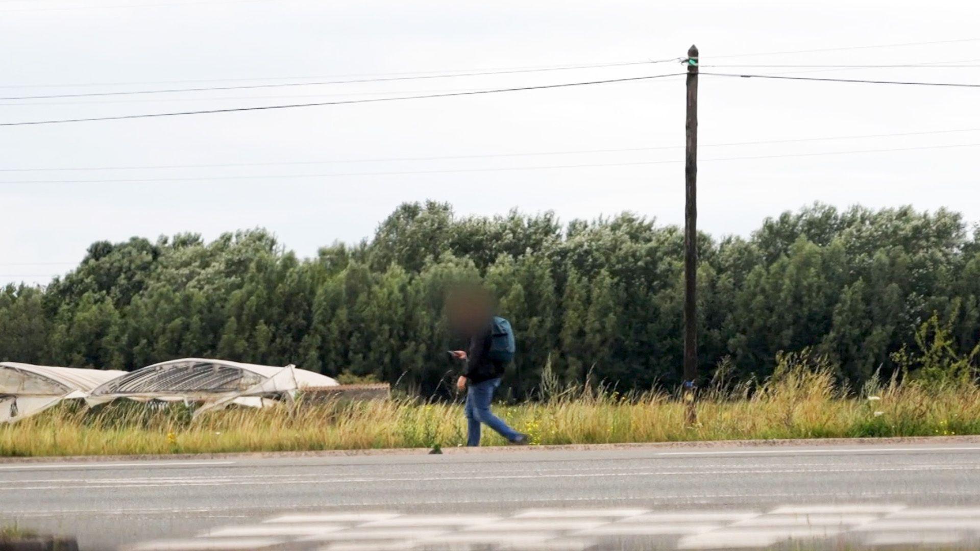 Man walks along an empty road with a wood in the background.