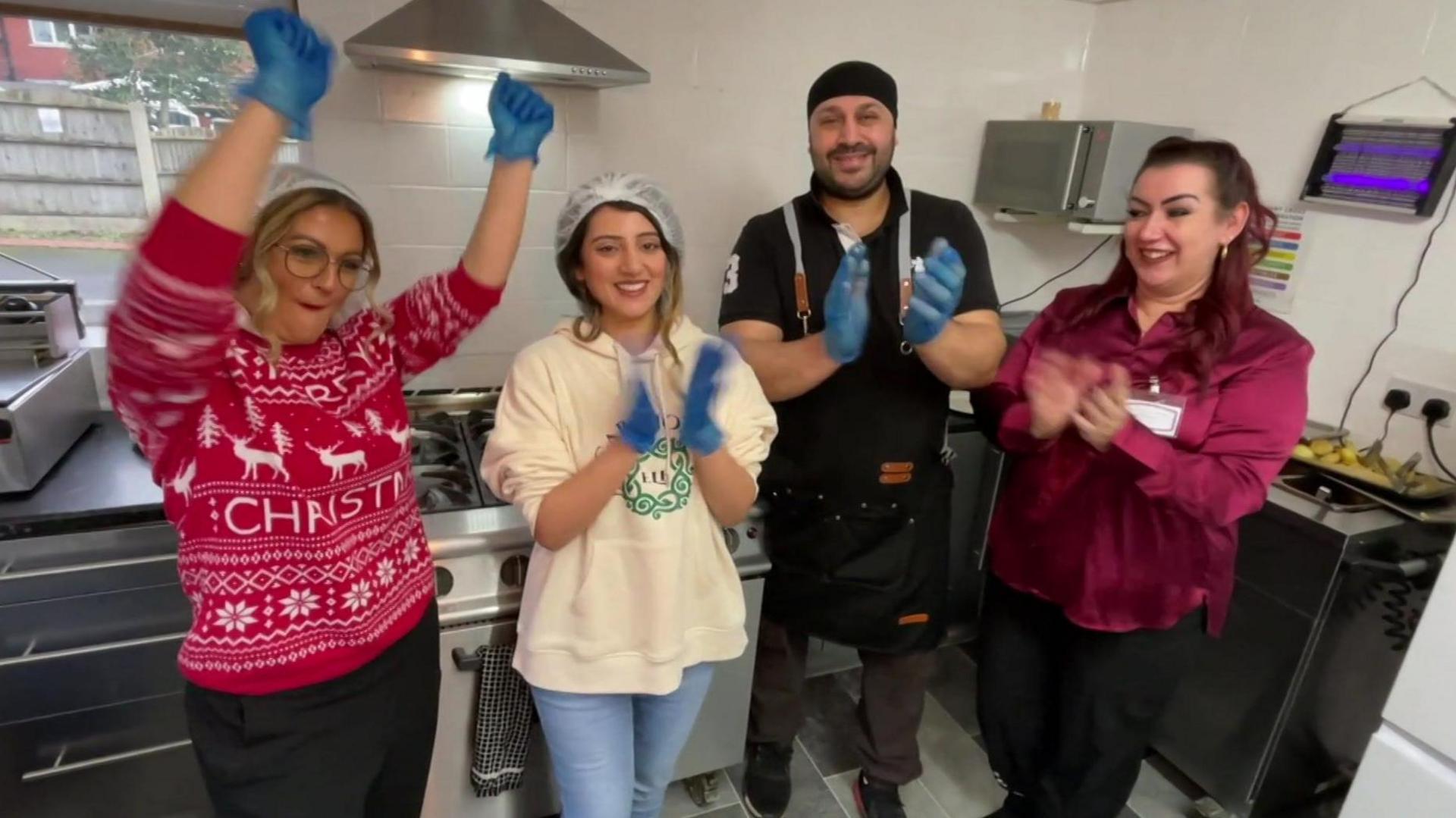 A woman wearing a Christmas jumper and dark trousers has her hands raised in the air, with Farrah Amjad standing next to her clapping while wearing a 'care home elf' hoodie and jeans. A man wearing a black apron, dark coloured top and brown trousers is also clapping his hands, along with a member of staff from the care home, wearing a silky cerise blouse and dark trousers. They are all smiling while standing in the kitchen at the care home.