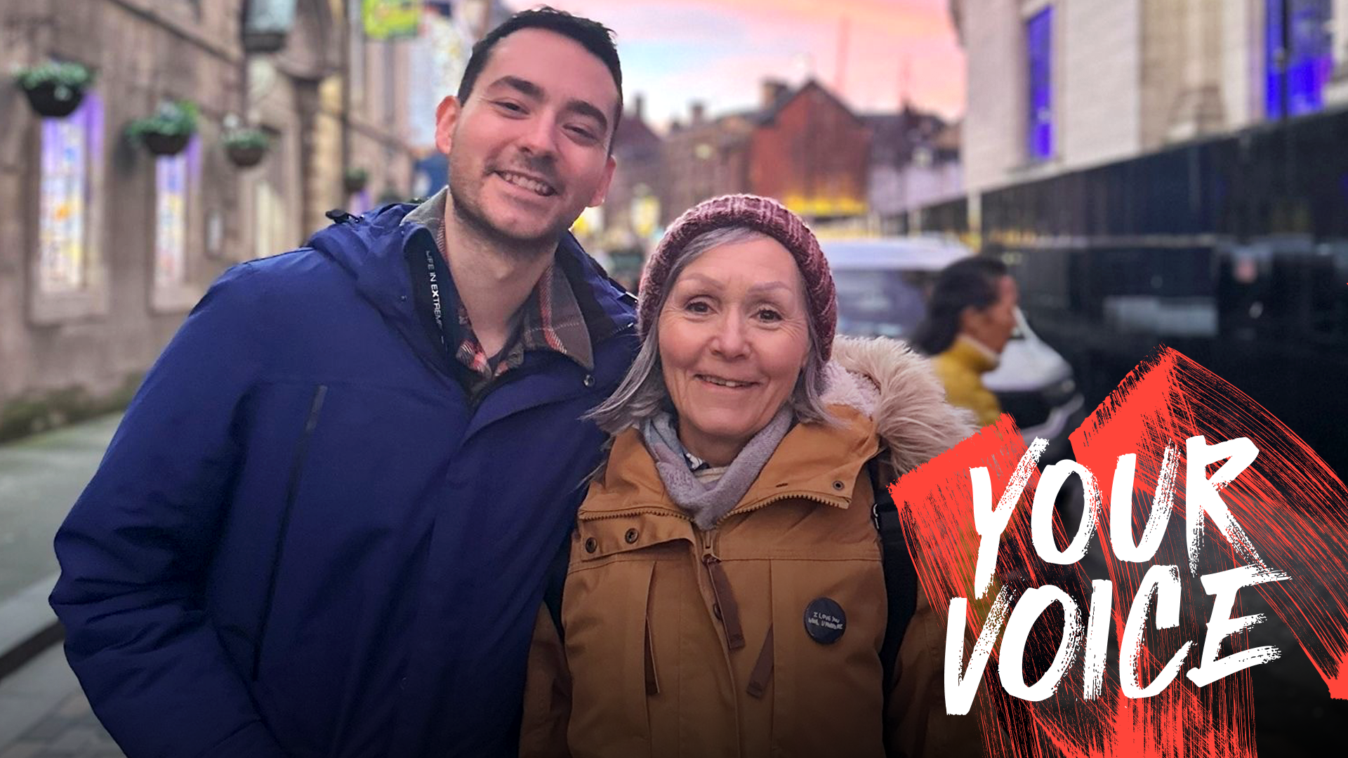 Deborah and her son Stuart smile while wearing coats - Stuart is wearing a royal blue one and hers is a burnt orange. They are standing in the street, with a pink sunset in the sky behind them. In the bottom right corner of the image is a white and red 'Your Voice' logo.