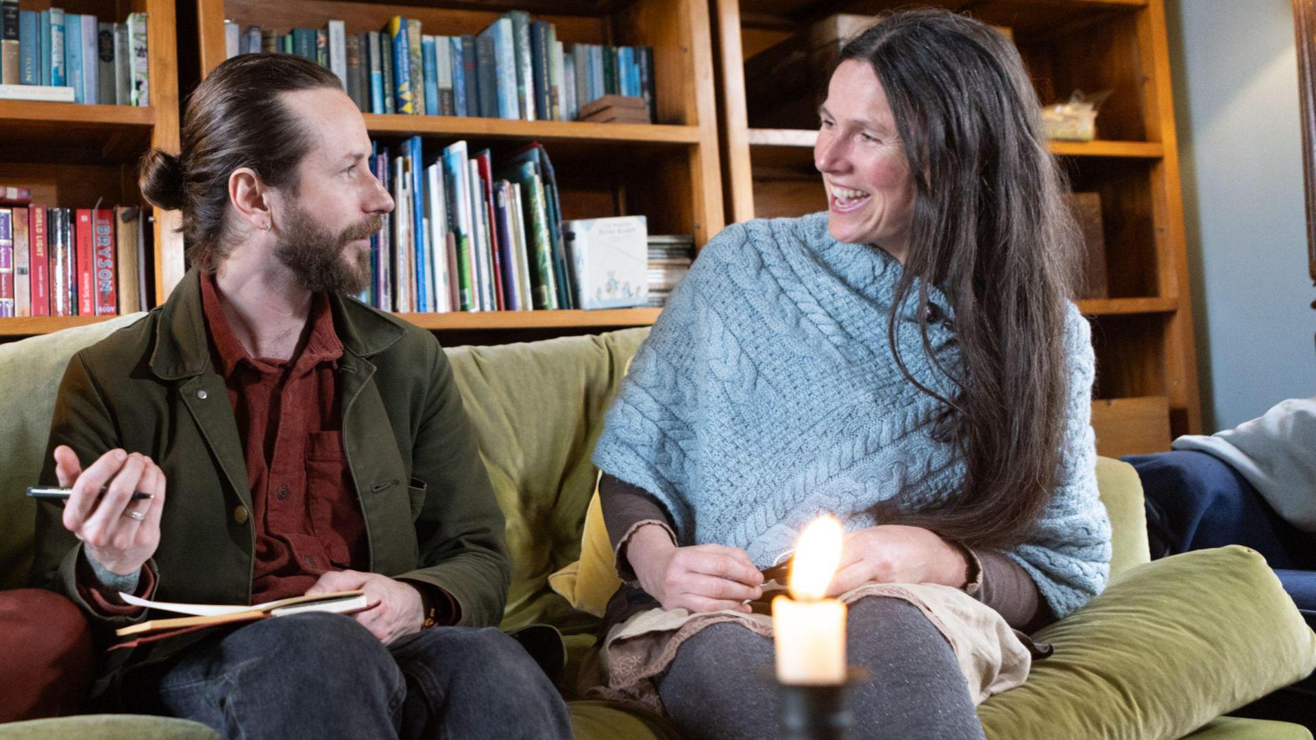 JP Worsfold and Jessie McMeekin. They are sat on a sofa talking to each other and smiling. Mr Worsfold is holding a notepad and pen. There are large shelves with books behind them.