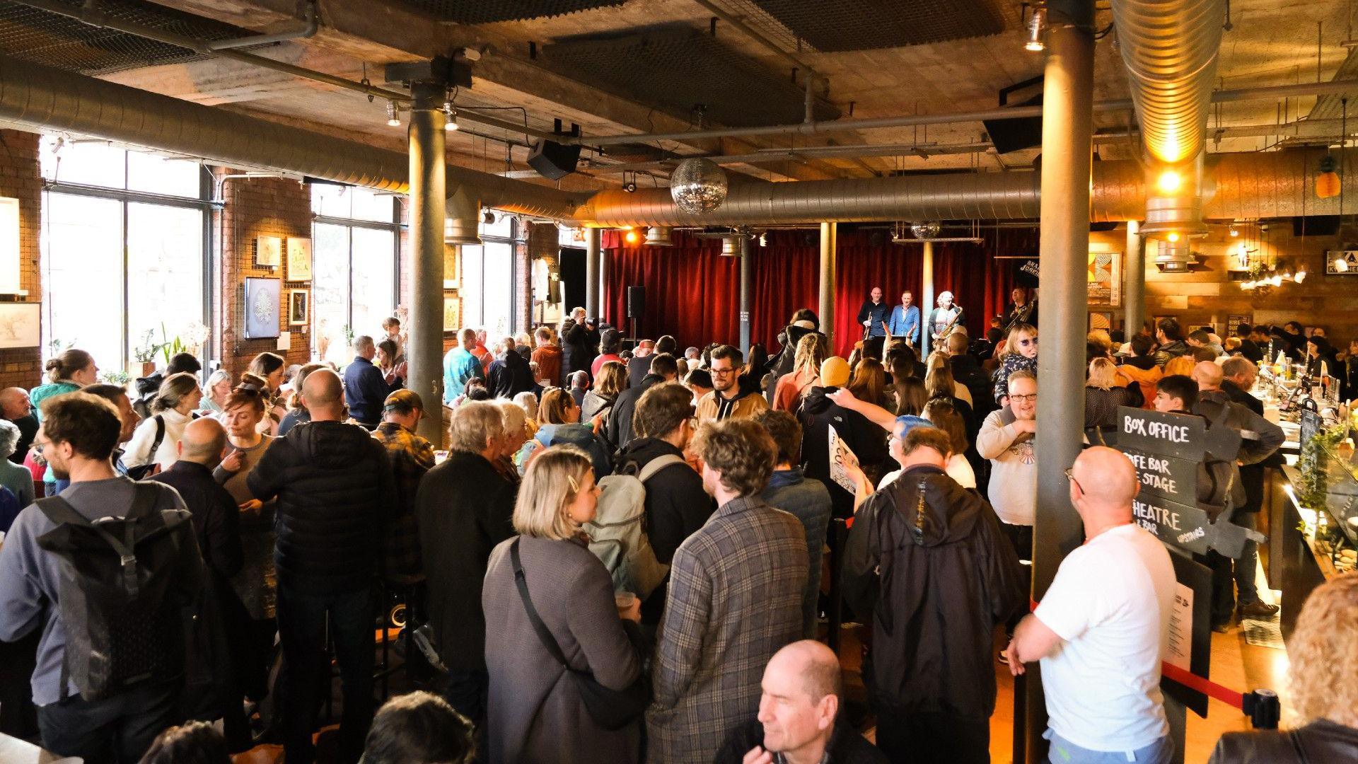 A crowd of people are seen chatting in an event space done out in an industrial style, with large, silver AC ducting on the ceiling. In the distance is a stage and a red curtain.