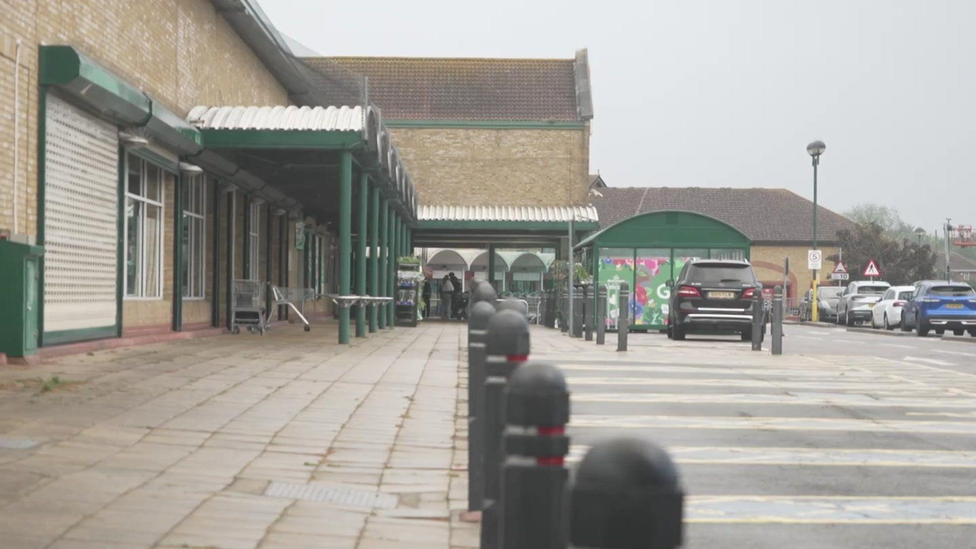 A brick supermarket with green arches is visible on the left behind black boulders, to the right are parking bays and some cars lined up