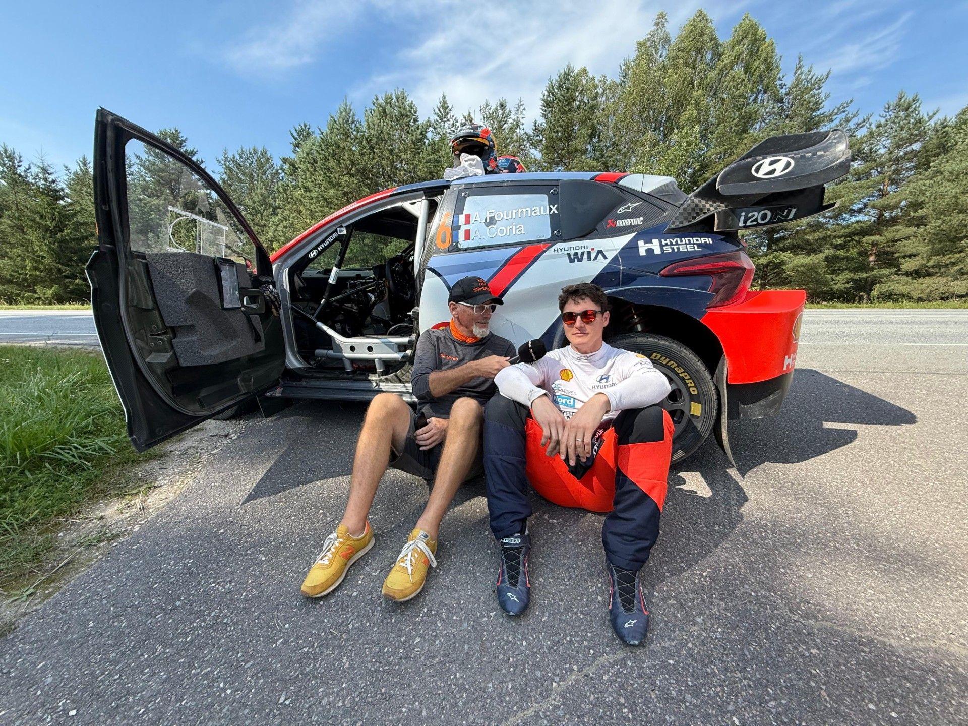 WRC commentator Colin Clark sitting down interviewing a male rally driver in front of his rally car