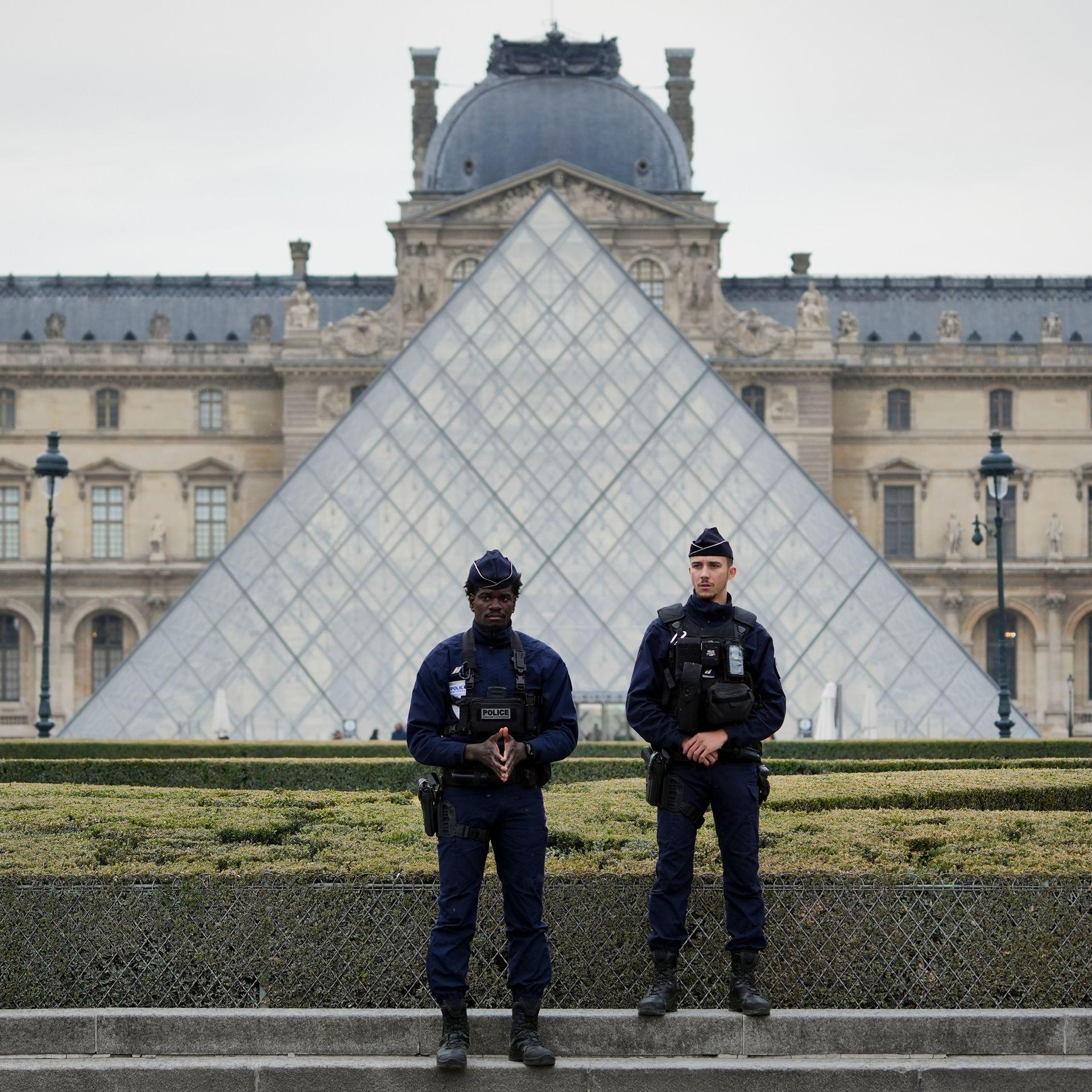 Two police officers in black uniforms stand guard in front of the iconic glass triangle of the Louvre museum in Paris.