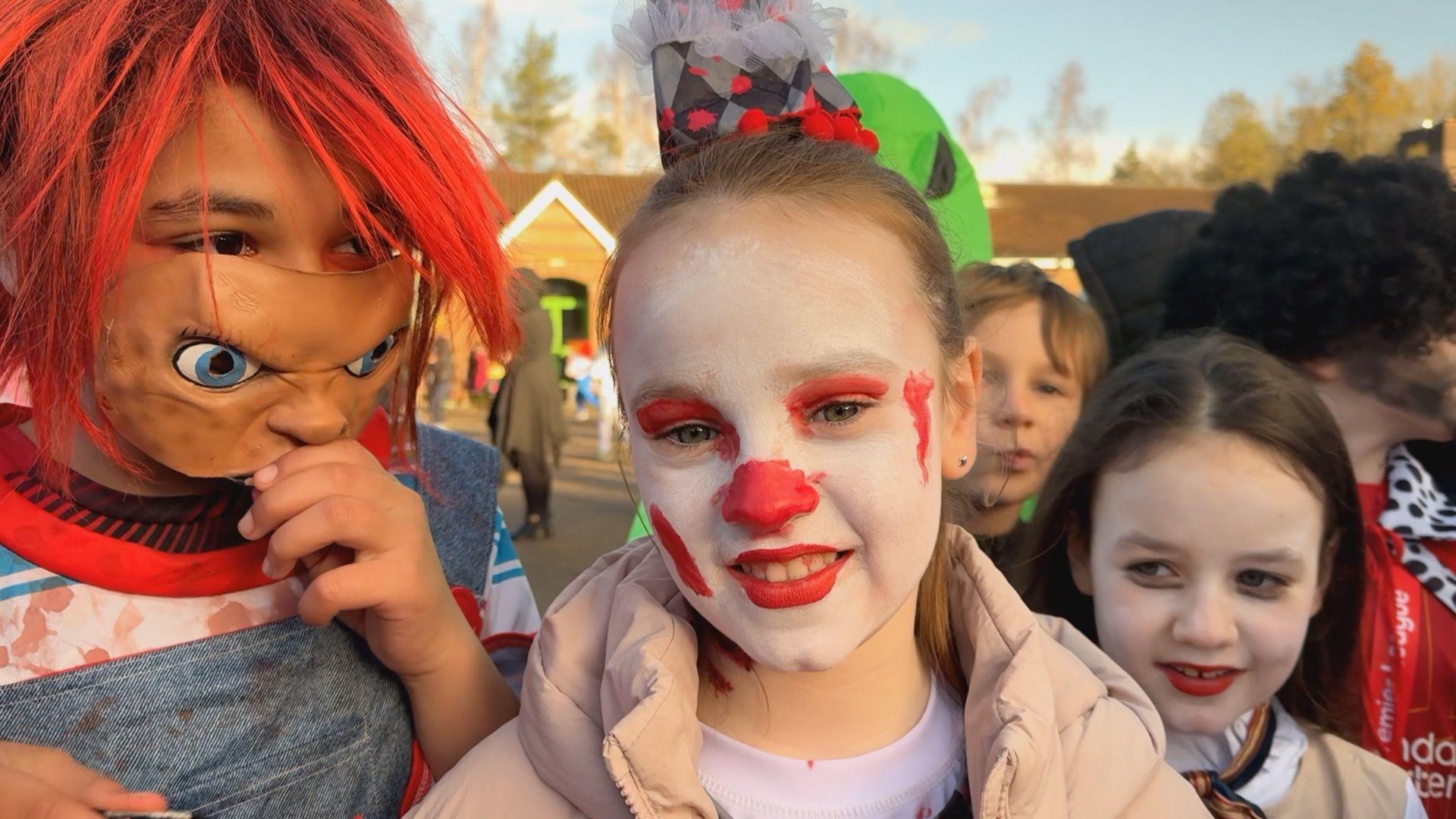 Evelyn is a schoolgirl and has her dark blonde hair pulled up on top of her head, she has white and red clown make up and is wearing a puffy pink coat over her costume. To her right is a child with a red wig and a brown mask wearing a pair of "bloodstained" dungarees.