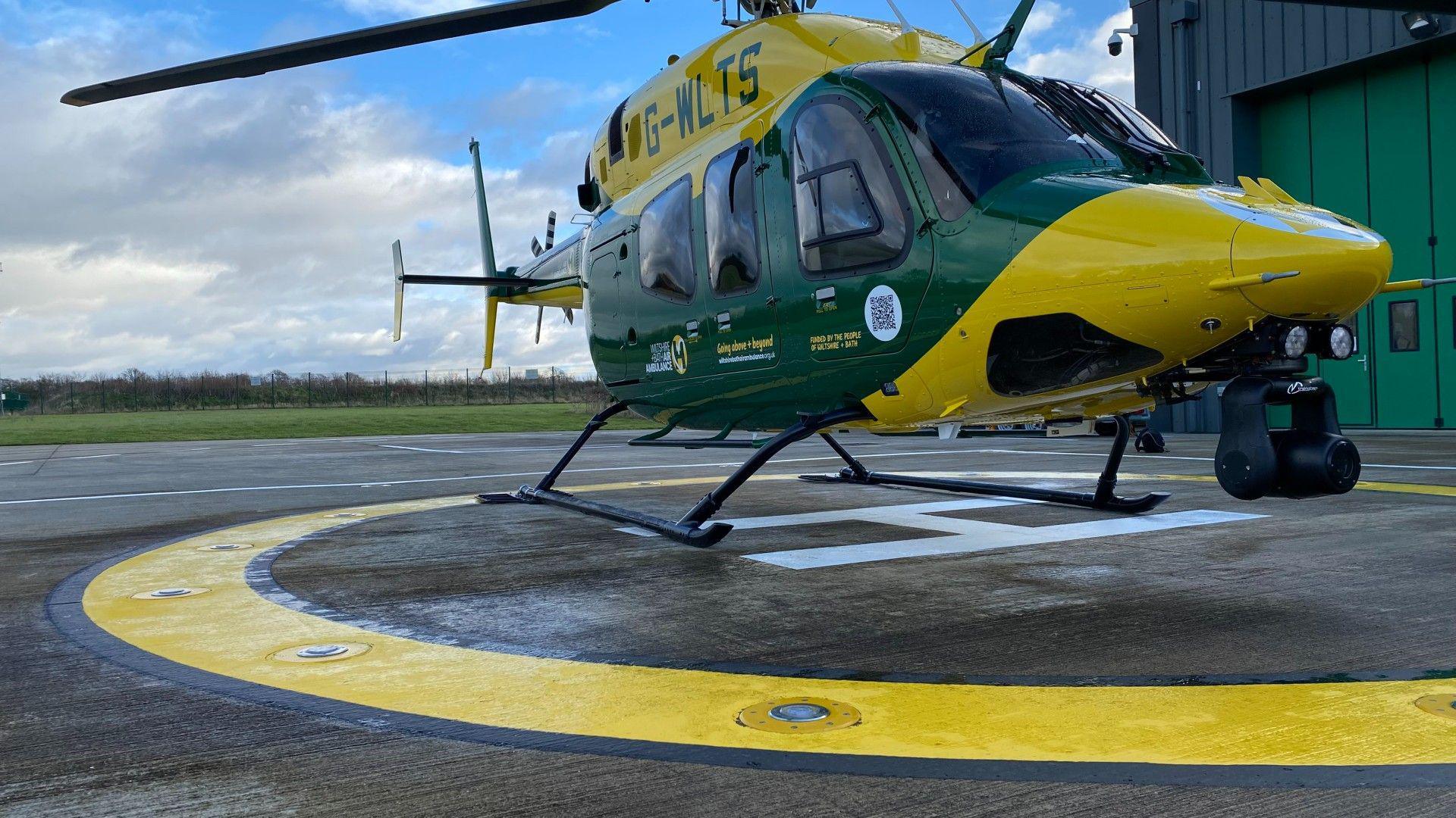 A yellow and green air ambulance on a helipad, next to a hangar on a cloudy day