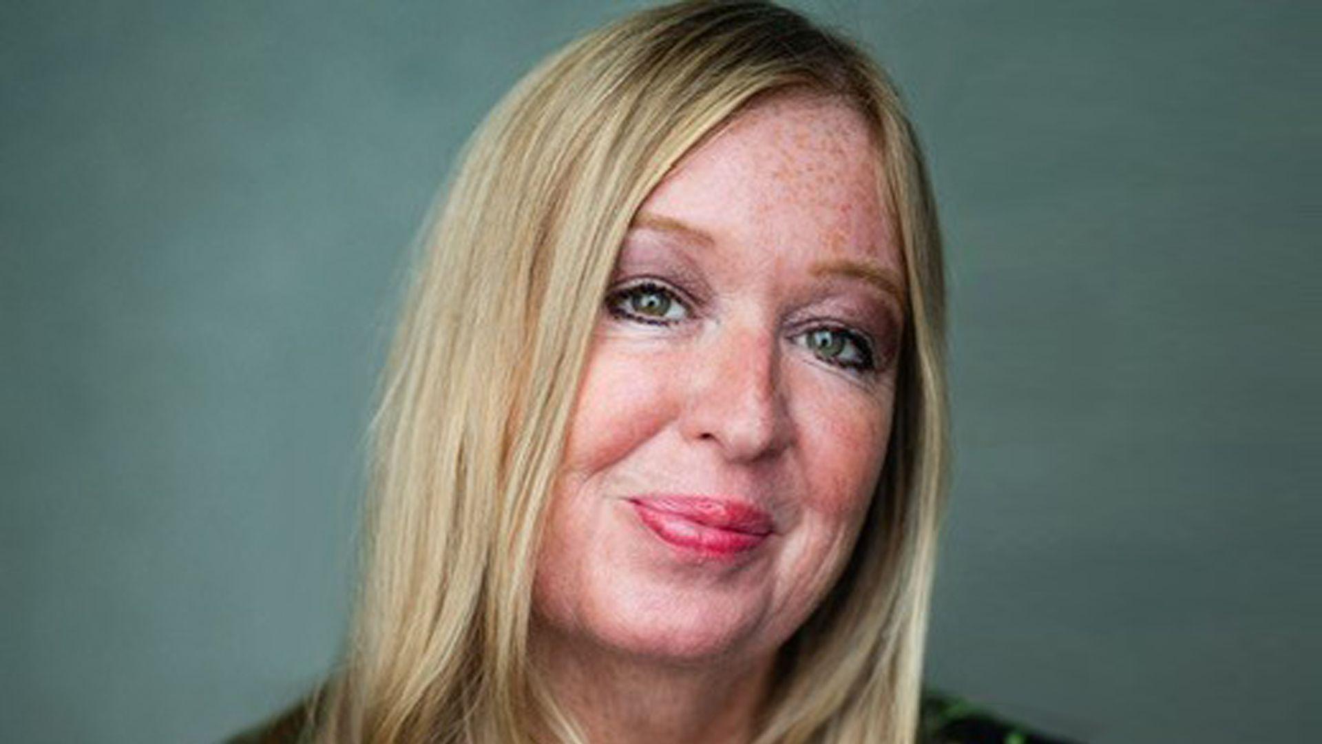 Debbie has long blonde hair and is smiling at the camera. She is in front of a dark green backdrop.