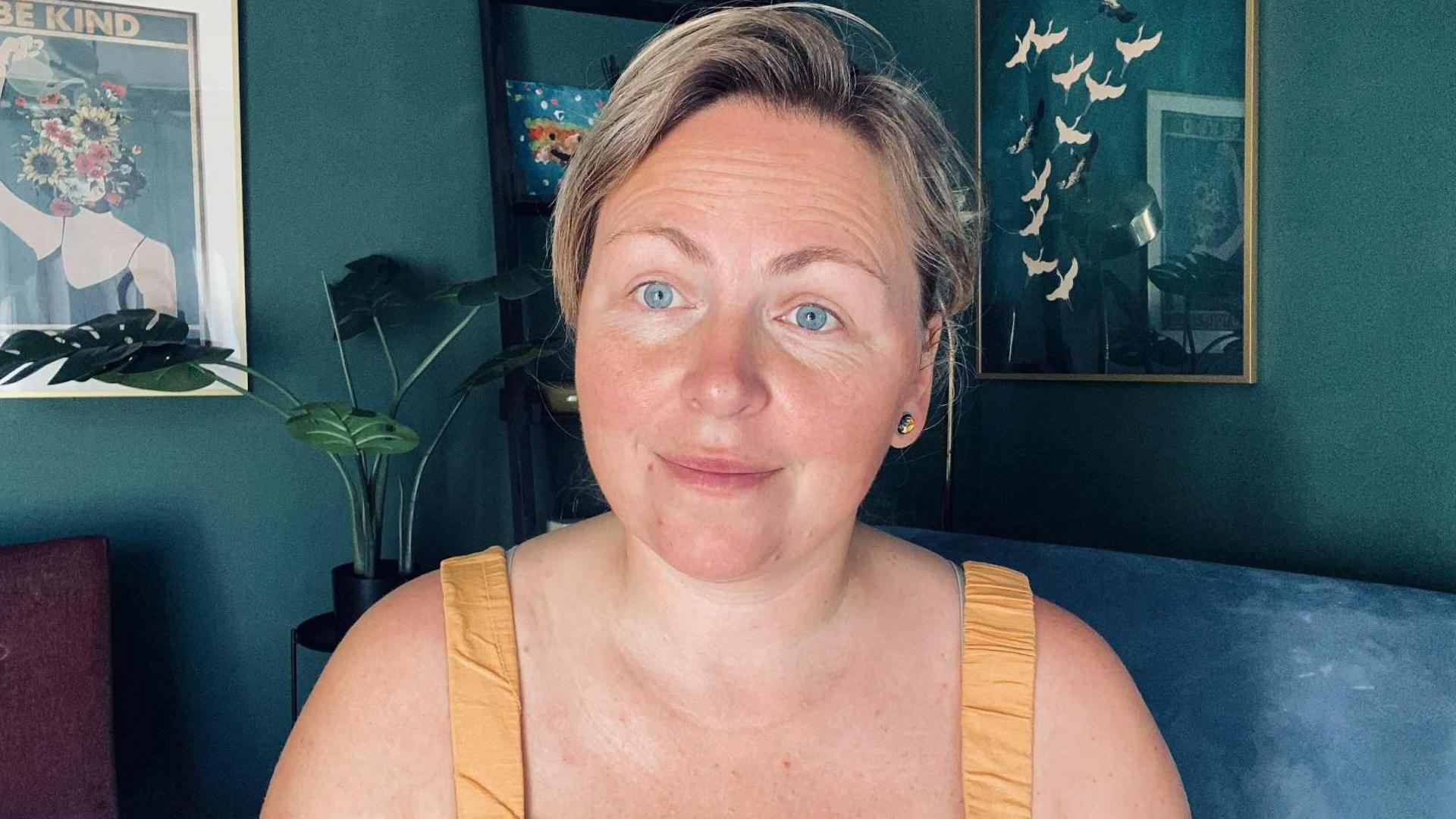 Patti sits in a teal living room with a plant and pictures behind her. She is smiling slightly and wears a strappy yellow top.