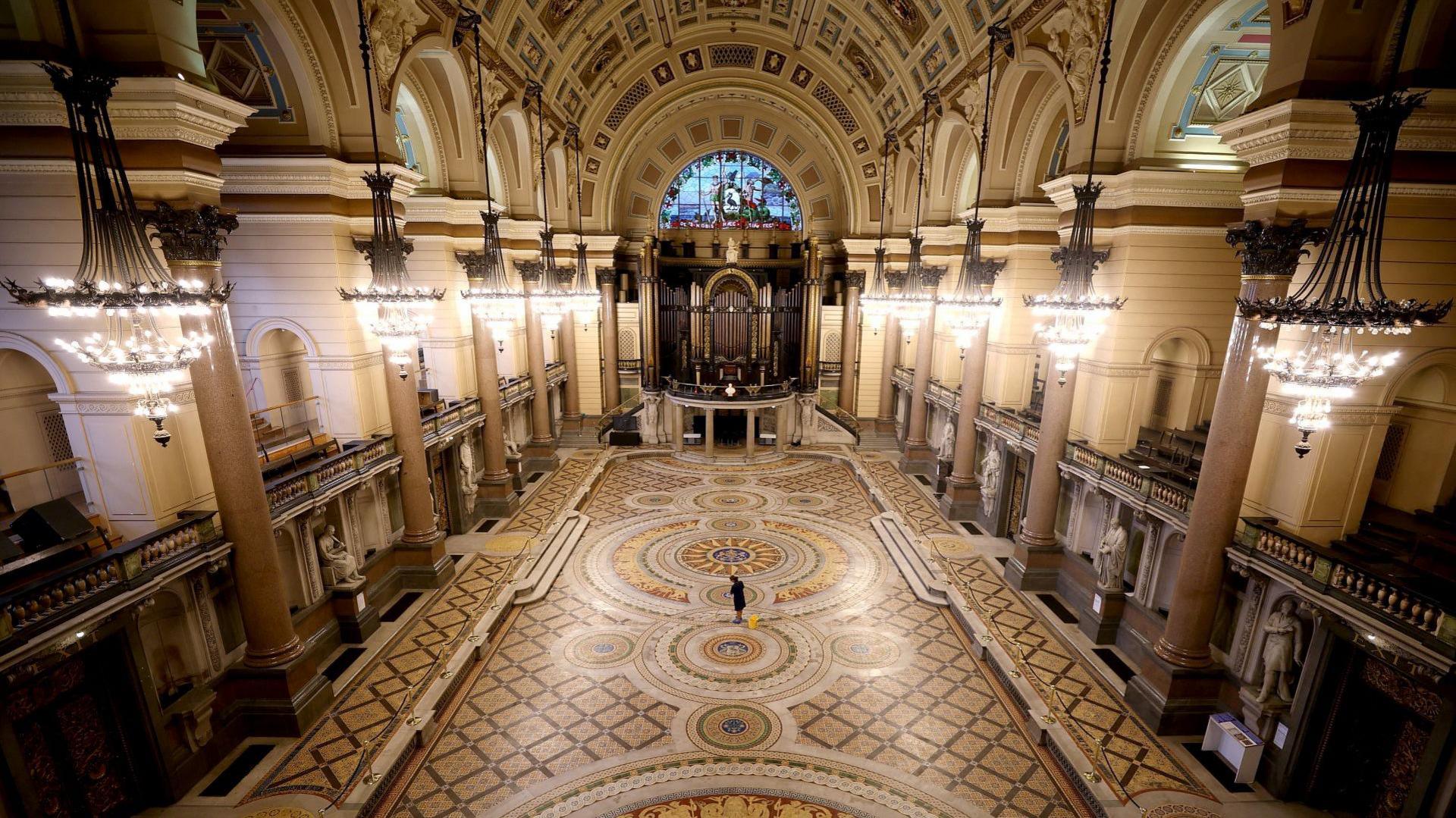 Interior of huge hall of St George's showing ornate multi-coloured mosaic floor and classical columns with ceiling arched above. Chandelier lamps hang from the ceiling.