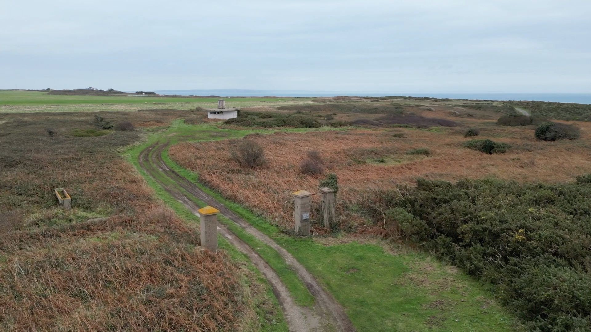 Aerial view of site of Lager Sylt camp in Alderney. A grassy and gorsey, windswept island landscape with a path and the remains of some concrete gate posts.