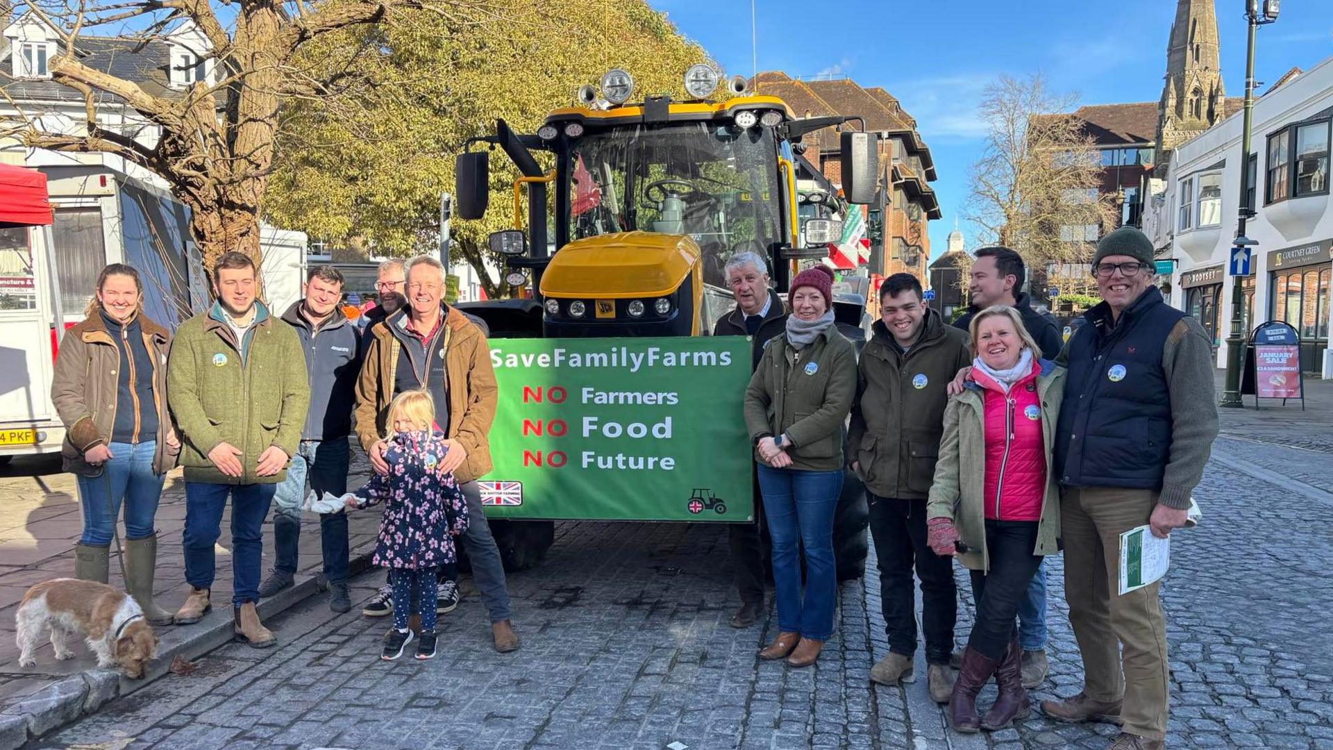 Roughly a dozen people stood in front of a tractor which is parked in a street