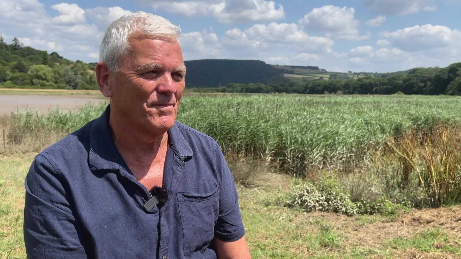 Philip is sitting in front of an area of wetland with reeds and grass under a blue sky. He has grey hair and is wearing a blue shirt.
