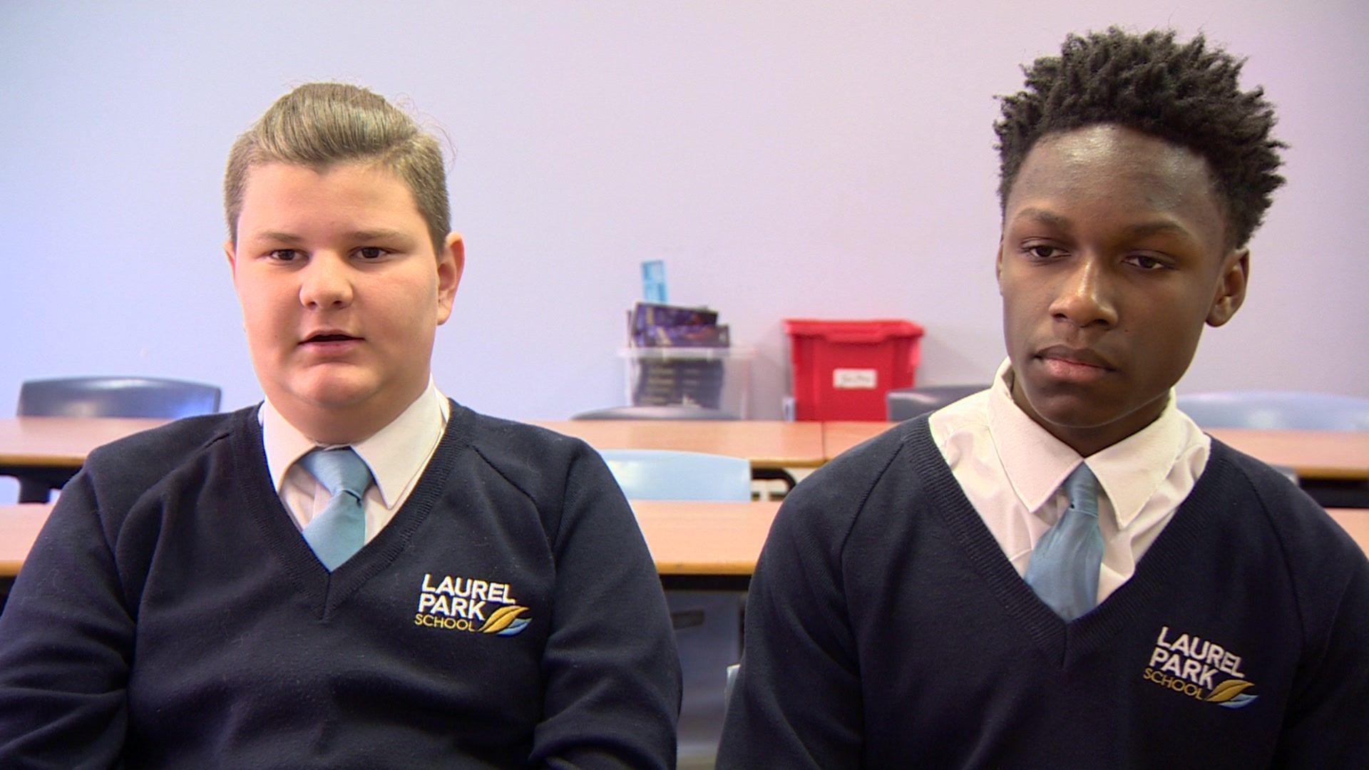 Two teenage boys in school uniform sit in a classroom with desks behind them