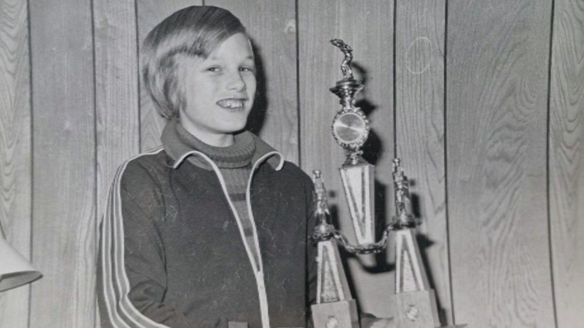 Adam is photographed as a child holding a table tennis trophy in this black-and-white image. He is smiling at the camera and is wearing a roll neck jumper and a sports jacket with stripes on the side. 
