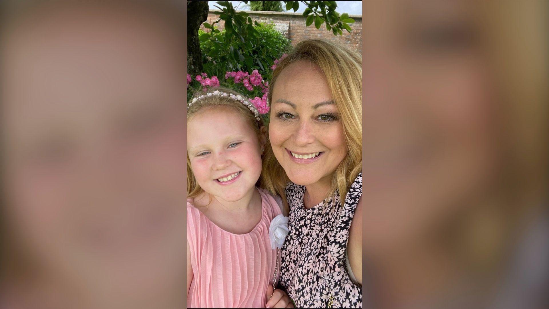 Michelle and her daughter Connie lean towards each other as they smile into the camera. Michelle has shoulder length blonde hair and wears a dark sleeveless top with a small white all-over flower pattern. Connie is wearing a pale pink pleated top and a white hairband holding back her blonde hair