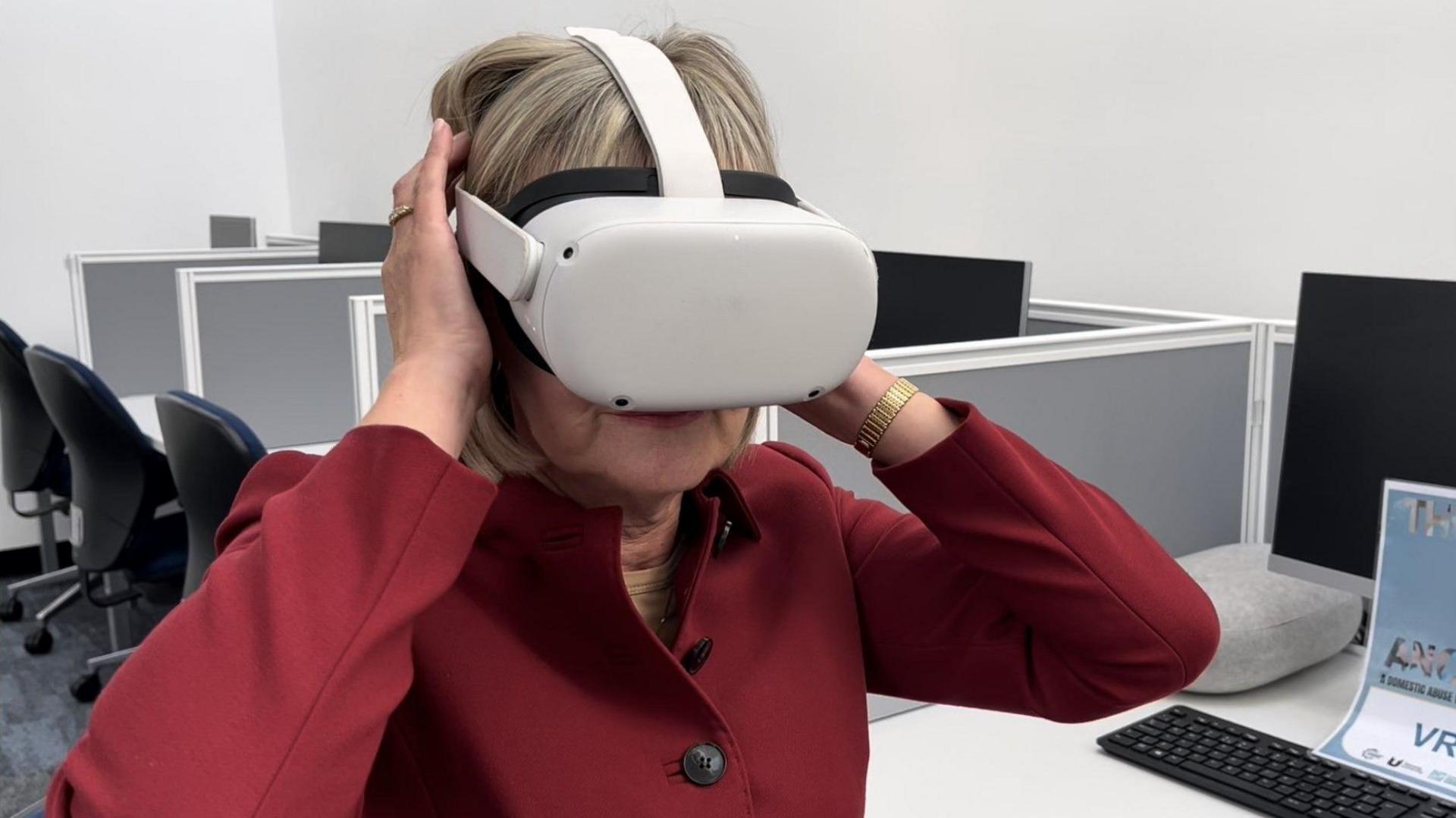 Durham Police and Crime Commissioner, Joy Allen, dressed in plum red wearing a white virtual reality headset with the strap over the top and side of her head, and a large electrical pack over her eyes. Her hands are supporting the strap.