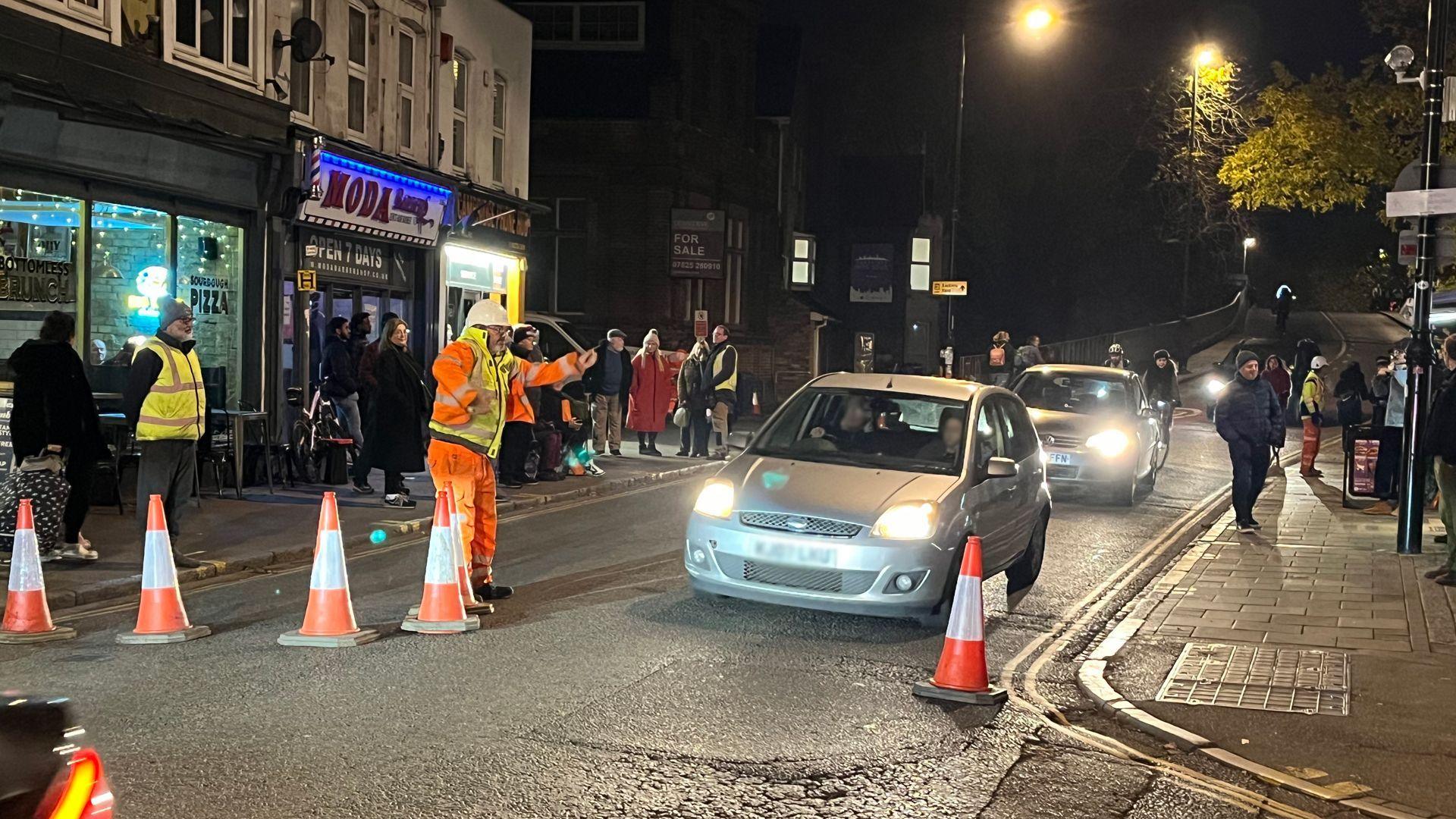 A workman in an orange outfit is waving cars to come through across Mill Road bridge in Cambridge. Six cones are in place on the road. People can be seen standing on either side of the road. Mill Road bridge can be seen in the background