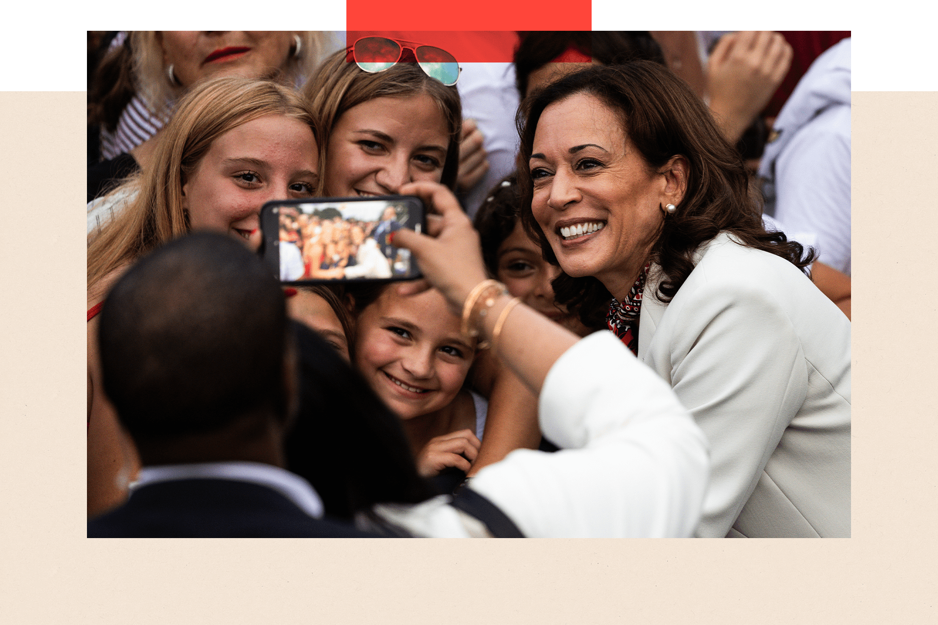 Kamala Harris in a white jacket having her photo taken on a phone. She is posing with a group of young women and what appears to be a child