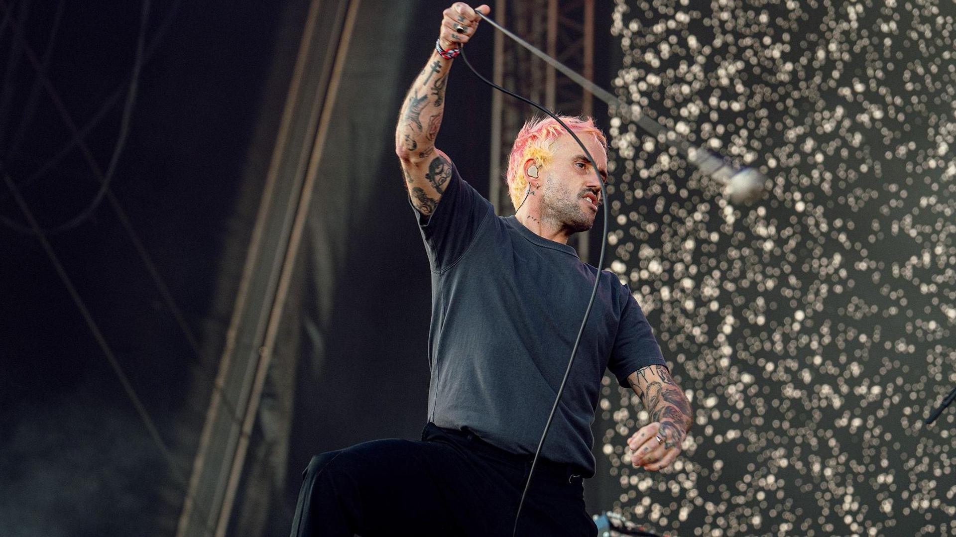 Joe Talbot from Idles twirls his microphone by the lead on stage. He has pink hair and is wearing a black t shirt and trousers