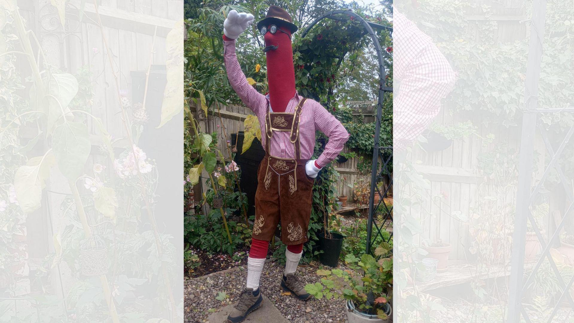 A man in a large red frankfurter costume, wearing traditional German dress, pictured in a garden.