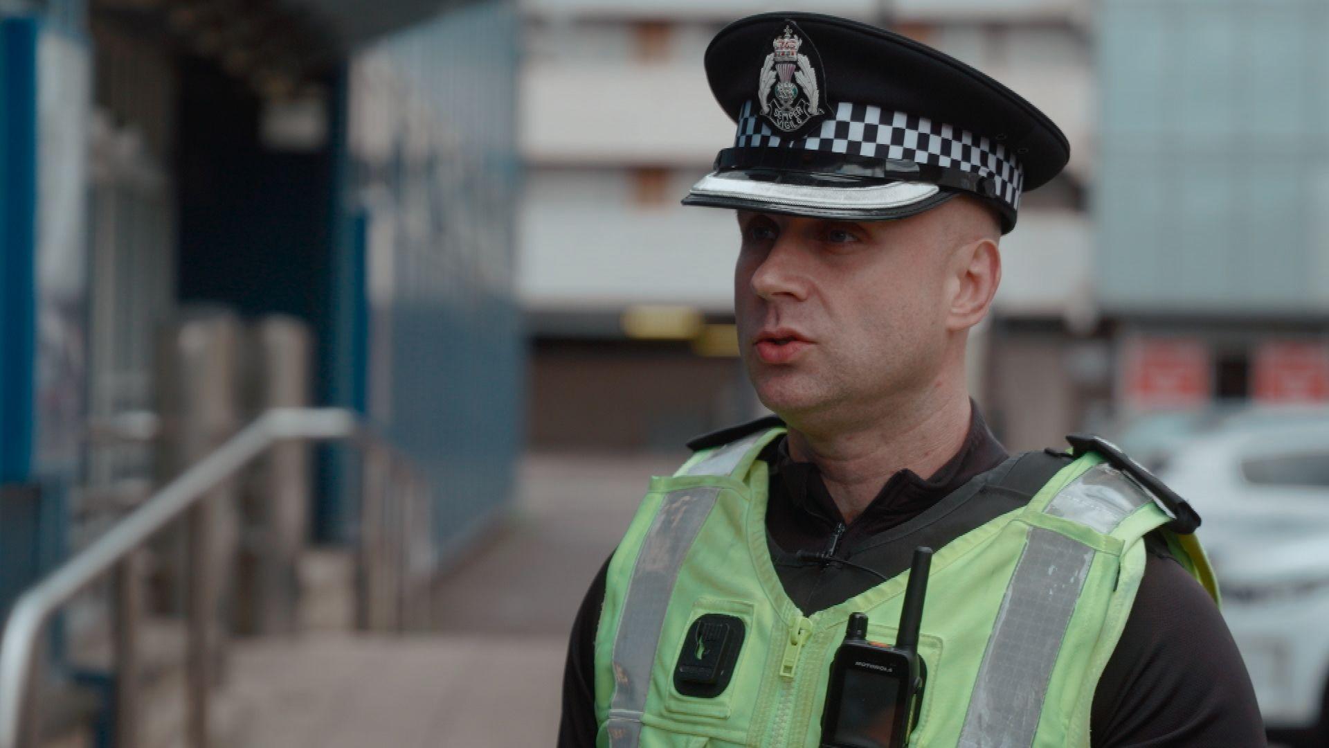 Ch Insp McManus wearing a high-visibility police uniform and checkered peaked cap, with a body-worn camera and radio attached to the vest. He is standing outdoors on a city street with buildings and parked vehicles blurred in the background.