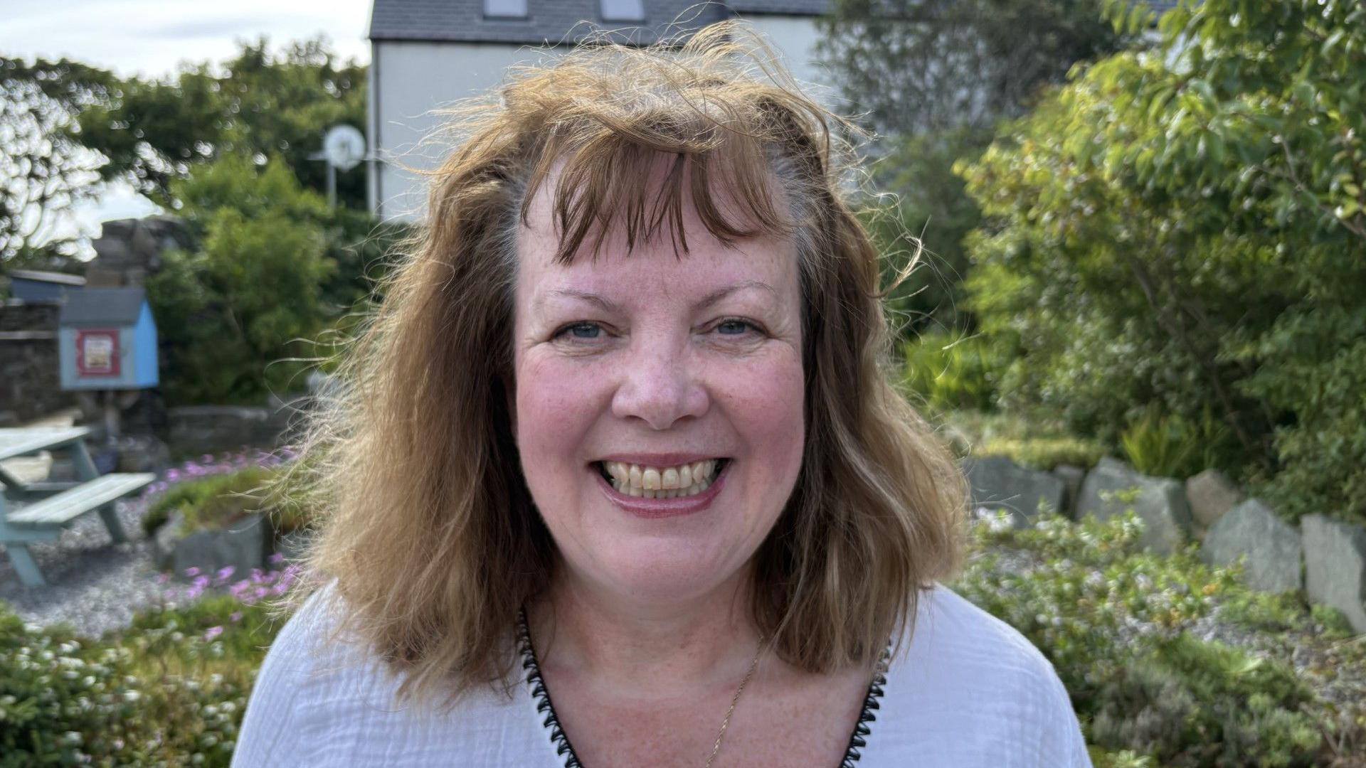 A middle-aged white woman smiling at the camera and standing in a public garden. She has brown hair and is wearing a white top. There's a picnic bench in the background and it's a sunny day.
