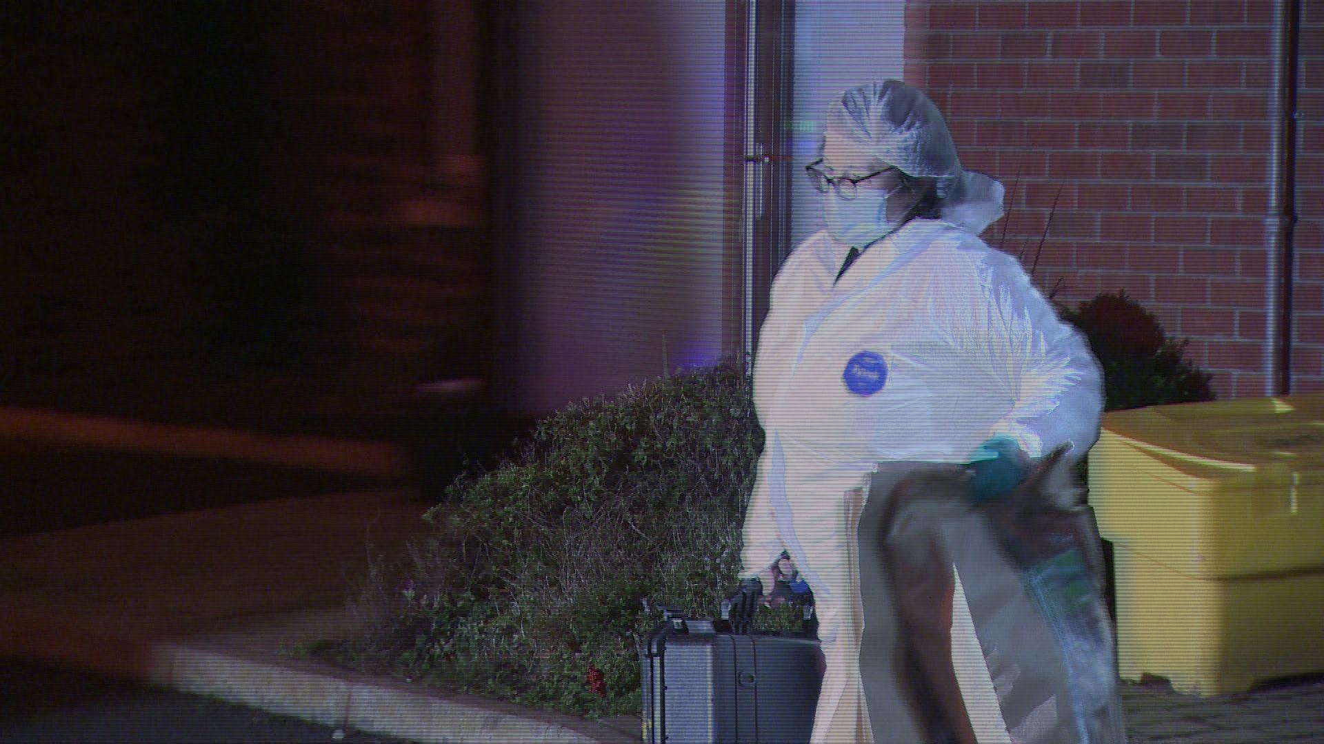 A female forensic officer in a white suit carries two large brown bags and a black box with a handle. She is wearing glasses and a face mask