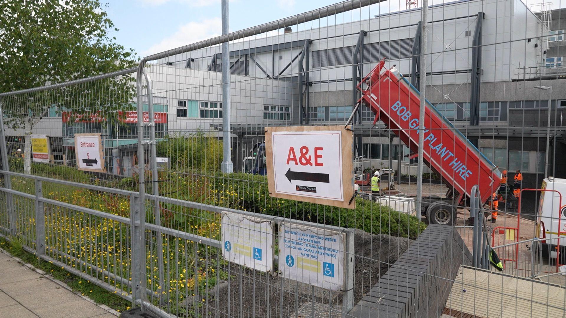 A metal hoarding with a temporary sign pointing to A&E at the Prince Charles Hospital. Through the metal railings a red tipper truck can be seen in front of the main hospital building.