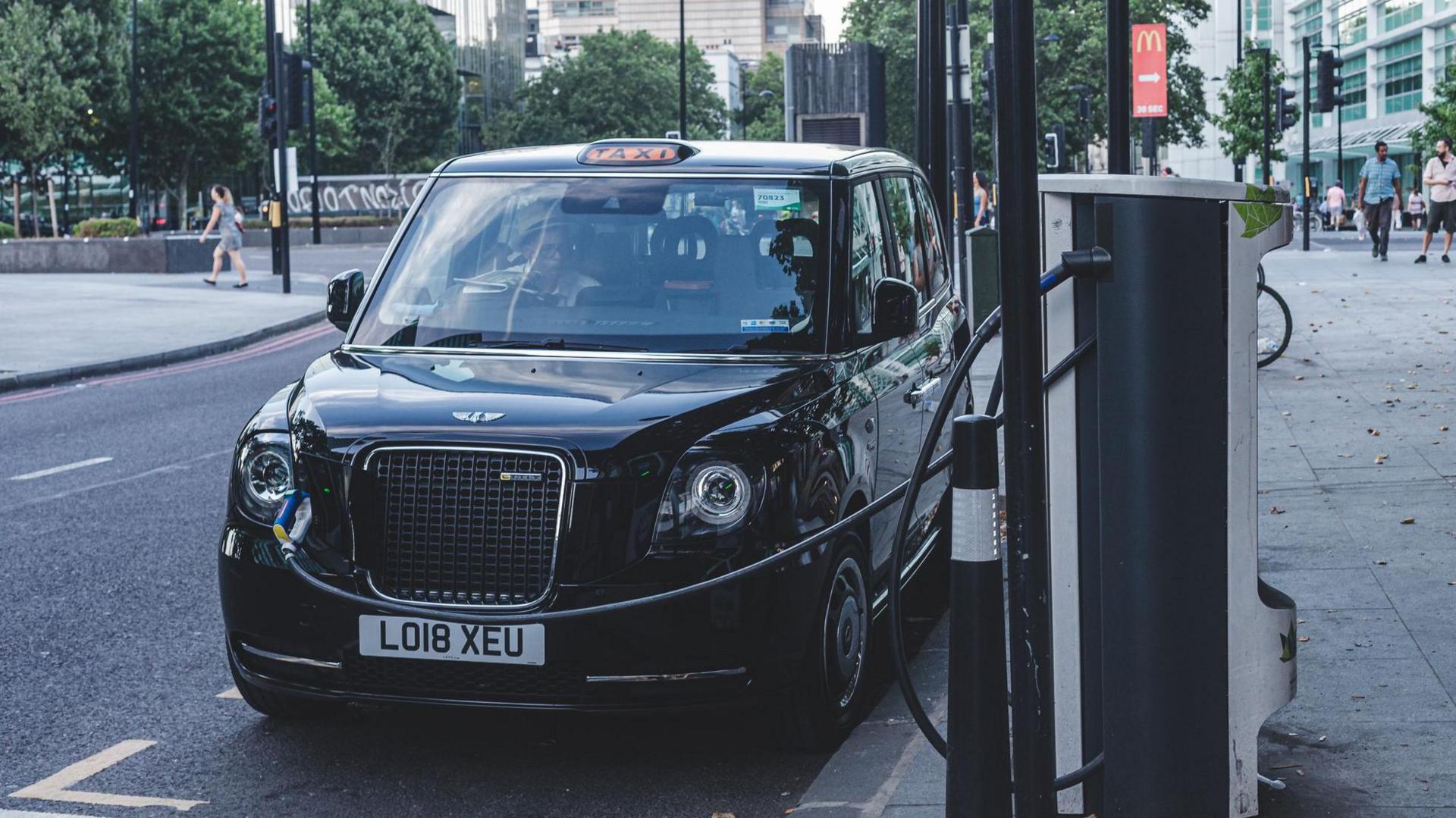 A black London taxi plugged into an electric charging point on a city street, with office buildings and pedestrians in the background.