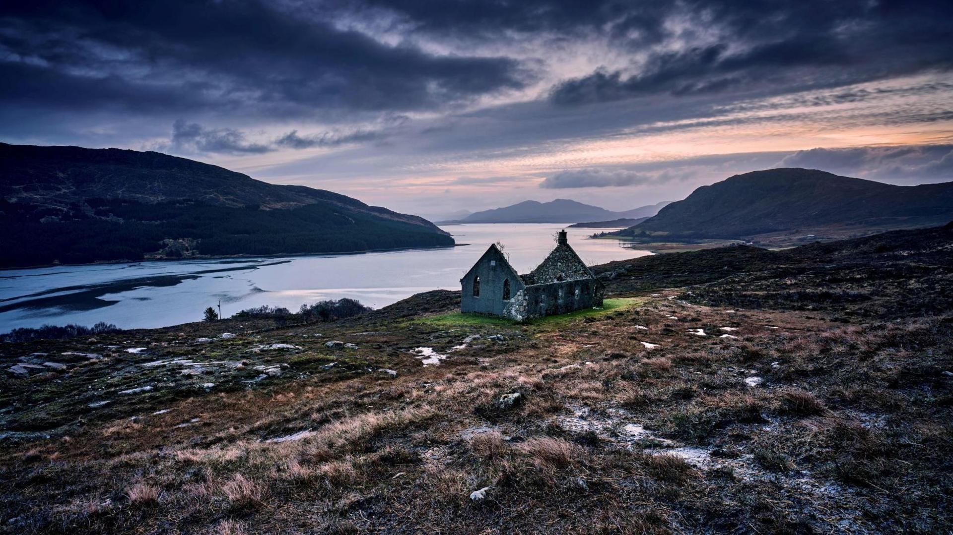 The church, which is roofless, is on a rugged moor above a loch. Hills and mountains rise from the loch. The sky is dark and atmospheric.
